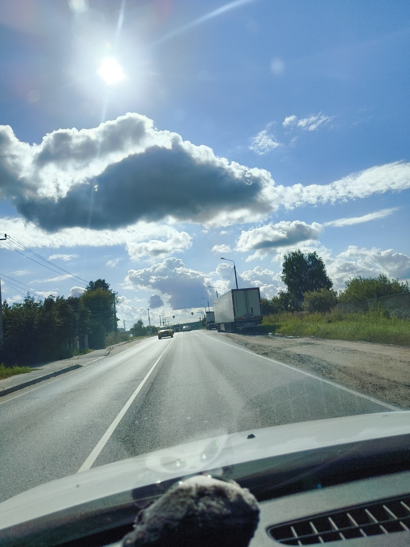 The image depicts a view from inside a vehicle traveling on a paved road with a white lane divider. The road runs through a semi-rural area with trees and grassy verges on either side. Several vehicles are visible on the road, including a yellow car in the foreground, followed by a white semi-trailer truck. In the distance, there are power lines and more trees. The sky is predominantly blue with scattered white and gray clouds. The sun is bright and visible in the upper left quadrant of the image, causing lens flare. The lower portion of the image shows the dashboard and windshield of the vehicle from which the photo was taken. The location is identified as Lyamtsino, Russia.