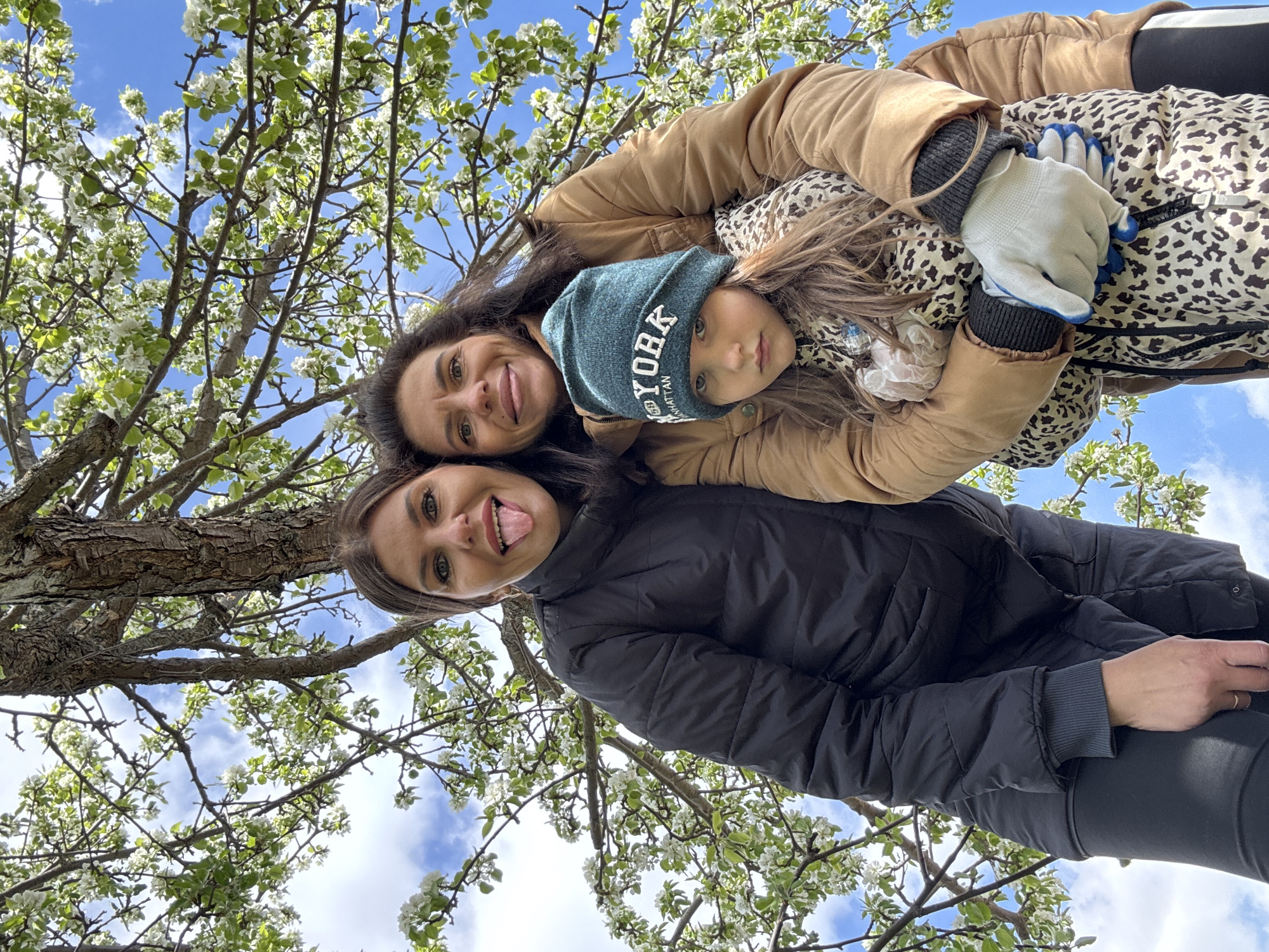 Three individuals are captured in an upward-angled photograph, likely taken in Dubovaya, Russia, given the query context. The background features a tree with white blossoms against a blue sky with scattered clouds.
A woman in a black puffer jacket and black leggings is in the foreground. She is sticking her tongue out. Above and partially behind her is another woman in a tan jacket, embracing a child wearing a blue "New York" beanie and a cheetah-print jacket. The child is also wearing work gloves. The people in the photograph are positioned under the tree.