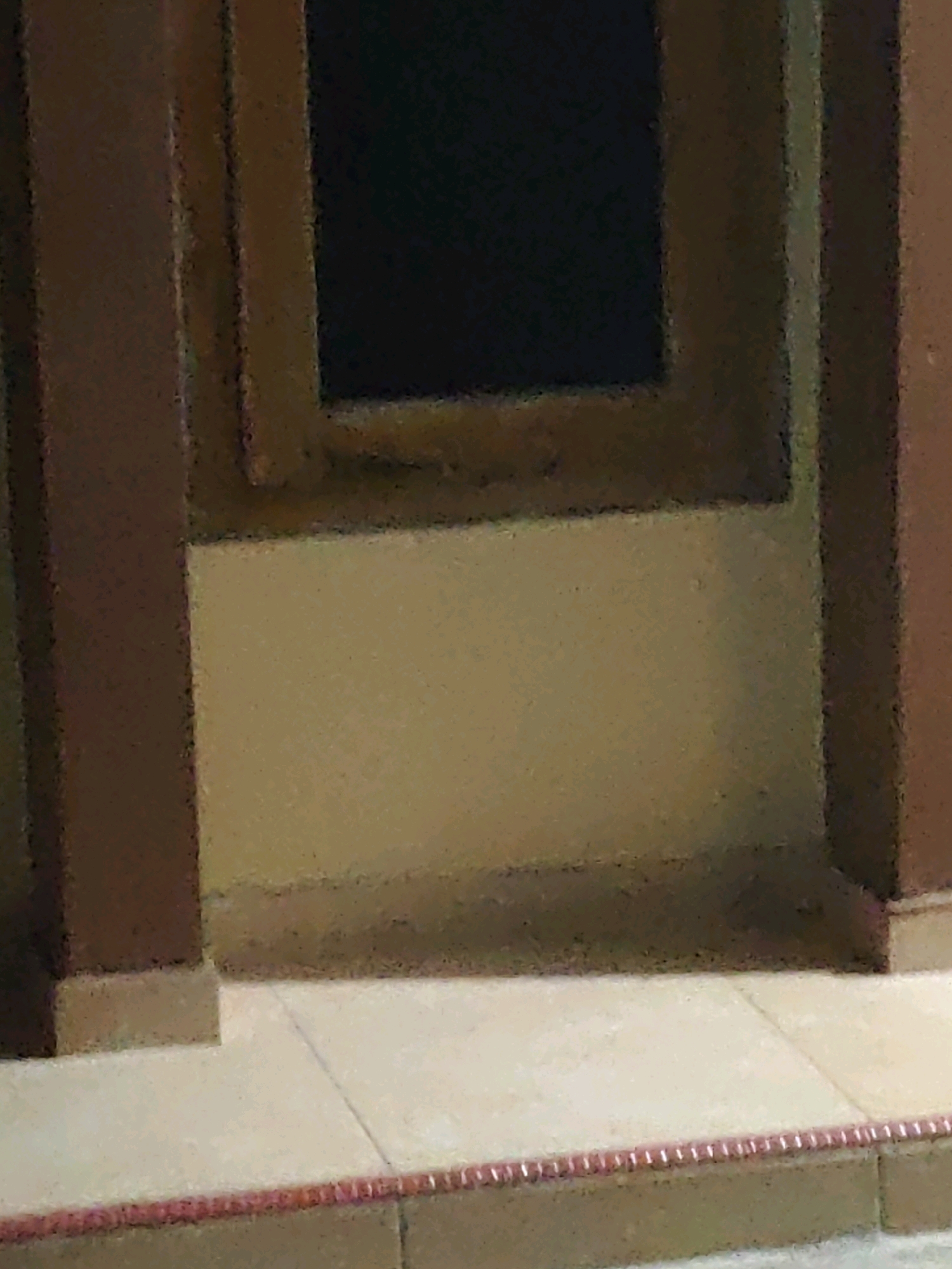 The image depicts a close-up view of a window frame and surrounding architectural elements. Two dark brown vertical posts frame the left and right sides of the scene. The window itself is dark, suggesting it may be closed or covered from the inside. Below the window, a textured, light beige wall is visible, with a distinct horizontal line separating it from a tiled surface at the bottom. The tiling is light-colored and appears to be arranged in square or rectangular sections, with a thin, decorative red and white border running along the edge of the foreground. The overall impression is that of an exterior architectural detail, possibly part of a building's facade. The location in Karang Sari, Indonesia, is not visually discernible from the provided image alone, as there are no specific geographical markers or context to indicate this location.