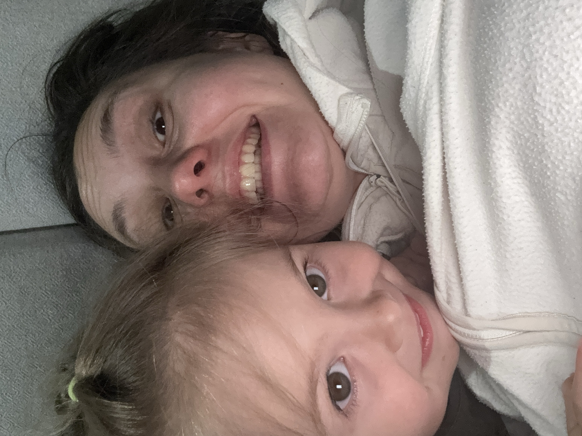 The image shows a selfie taken indoors, presumably at an elevated angle looking down. Two people are present: a woman and a young child. The woman has dark hair, fair skin, and is smiling. She is partially covered by a white, textured blanket or towel. The child, also fair-skinned, is positioned below the woman and is looking up at the camera. The background appears to be a gray upholstered surface, possibly a couch. There is no evidence to determine the location is in the city of Zelenogradsk, Russia.