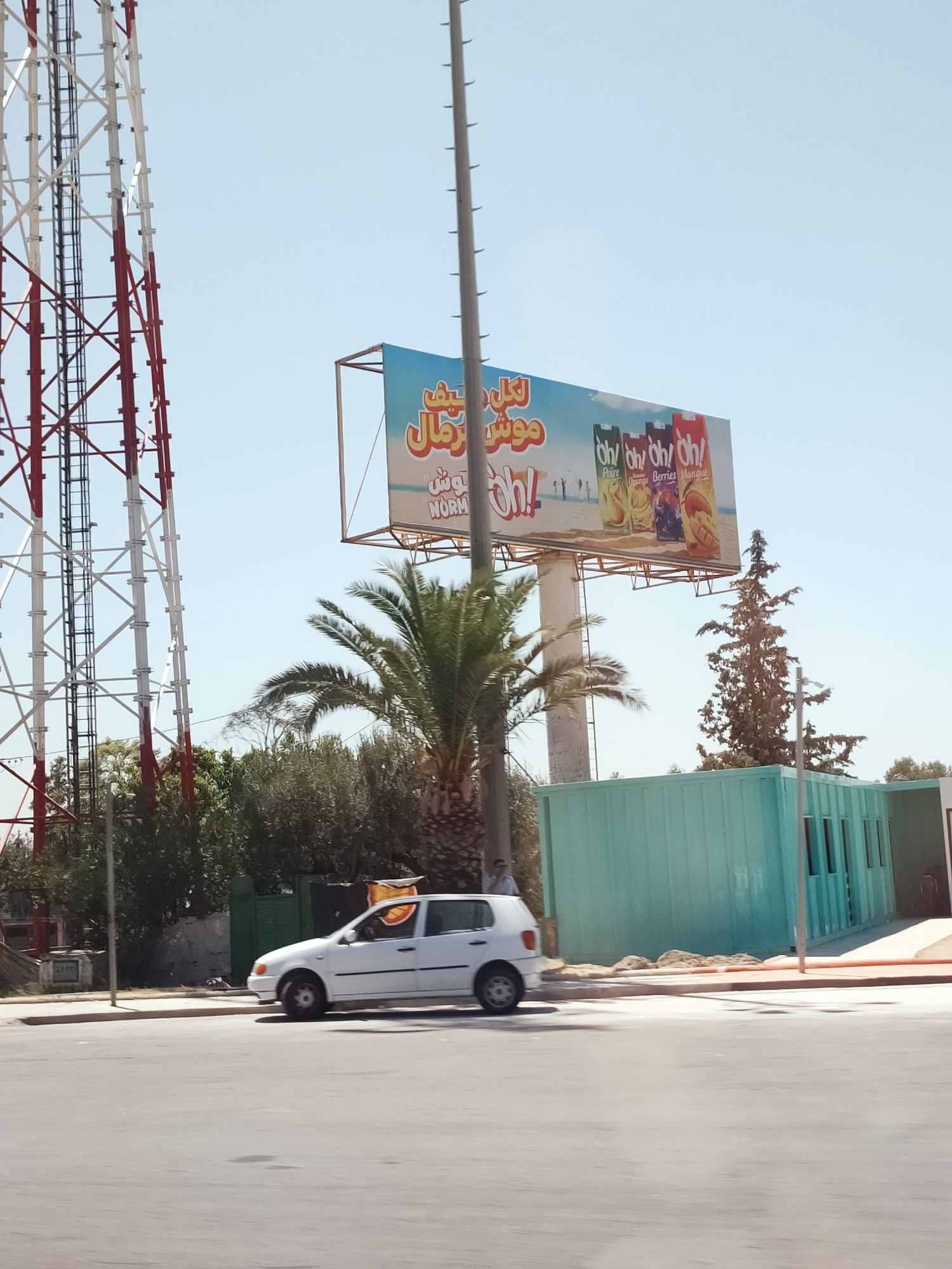 The image shows an outdoor scene with a large billboard for "Oh!" juice drinks. The billboard features images of four juice cartons with flavors such as "Poire," "Orange," "Berries," and "Mangue," set against a beach background with silhouetted figures. To the left of the billboard is a tall, red and white lattice tower, likely a telecommunications tower. In front of the billboard and tower, a white hatchback car is visible driving on a paved road. A palm tree and other green foliage are present in the foreground and midground. Behind the car and billboard is a teal-colored corrugated metal building with several windows. The scene appears to be in an urban or semi-urban environment under a clear blue sky. The location is specified as the city of Crétéville, Tunisia. There are no prominent interactions depicted between people or objects.