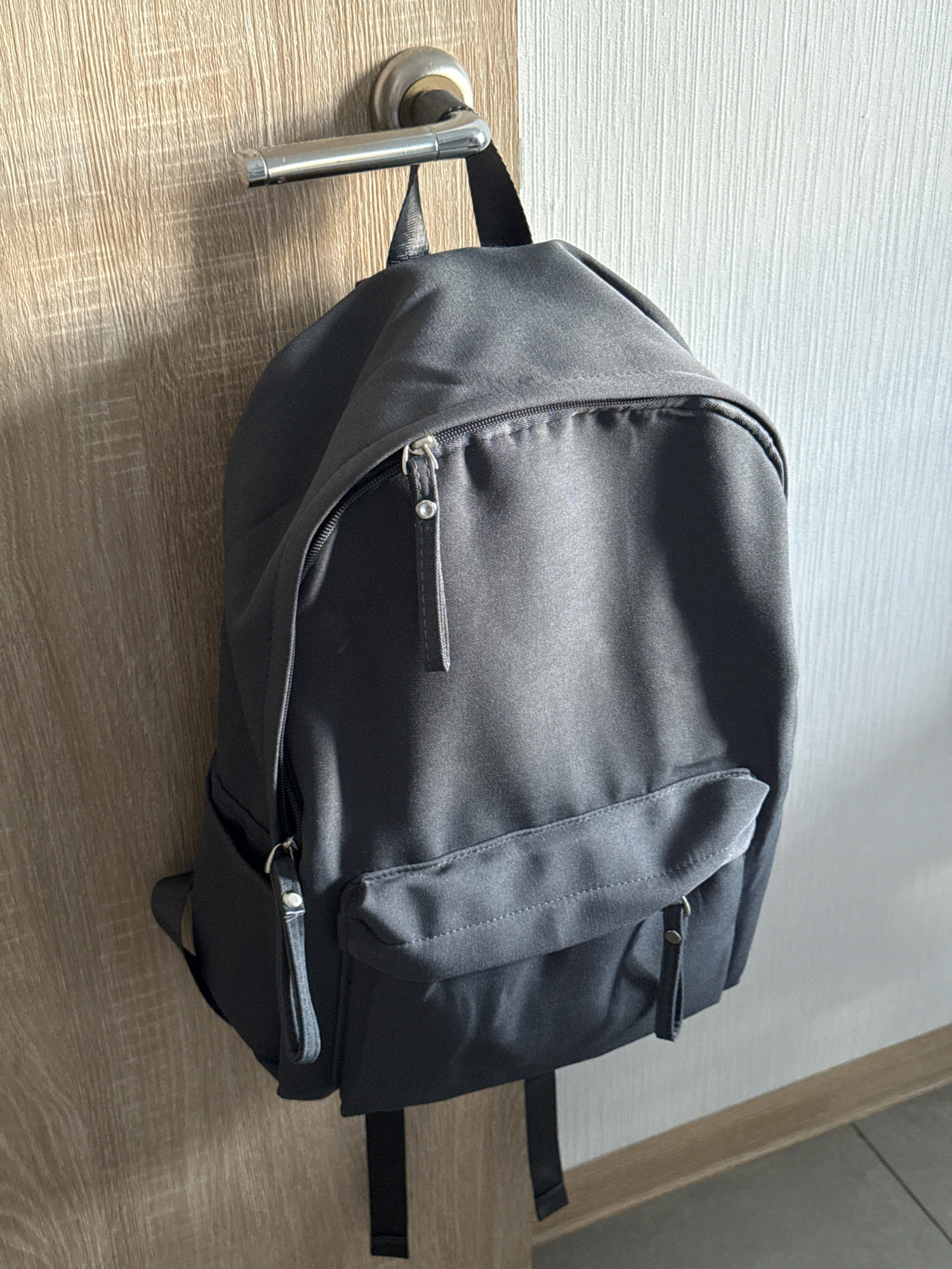 A dark gray backpack is hanging from a silver door handle on a light wood door. The backpack has a main zippered compartment, a front zippered pocket, and a flap pocket on the front. There are two black straps hanging down from the bottom of the backpack. The door is on the left side of the frame, and a textured white wall is on the right. The lighting suggests an indoor environment. The scene is consistent with a typical residential setting in Lyubertsy, Russia, where such doors and backpack storage are common.