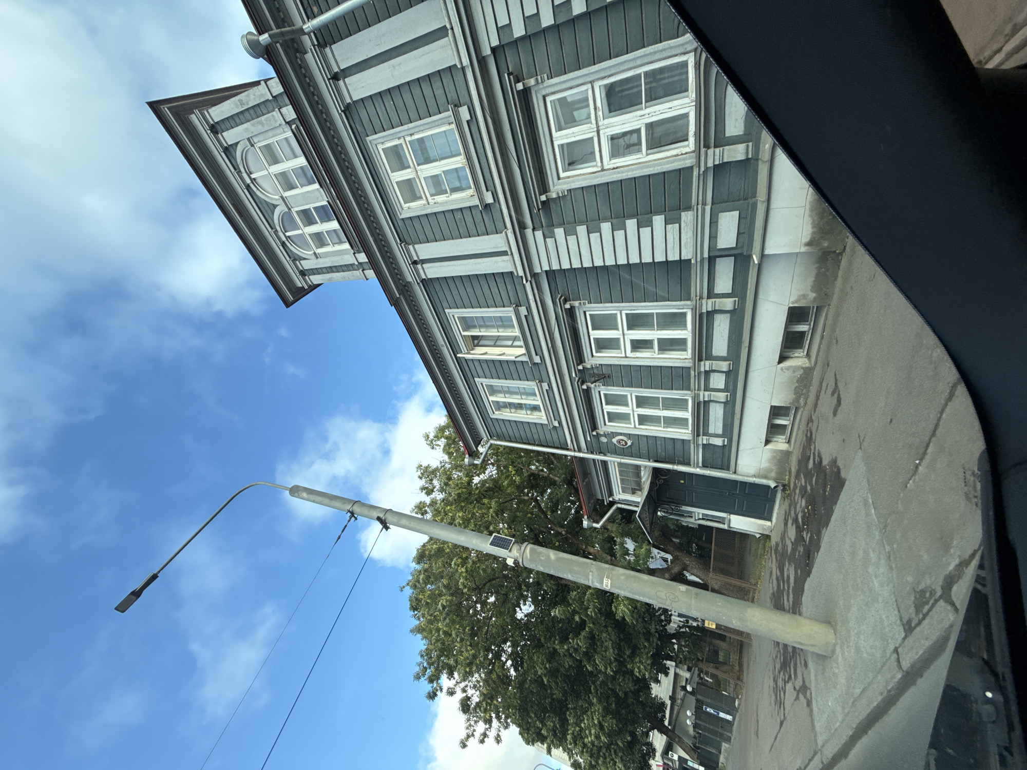 This image captures a low-angle view of a historic, two-story wooden building in Tallinn, Estonia, with a distinct teal-green facade and white trim. The building features numerous multi-paned windows, some of which are open, and an ornate mansard roof with arched dormer windows. A lamppost with a solar panel attached to it stands on a paved sidewalk in the foreground, leaning slightly. The street in front of the building appears wet, suggesting recent rain. Lush green trees are visible behind the lamppost, and the sky above is a vibrant blue with scattered white clouds. The perspective is from within a vehicle, as indicated by the dark interior element along the bottom right of the frame, suggesting the photograph was taken from a car window.