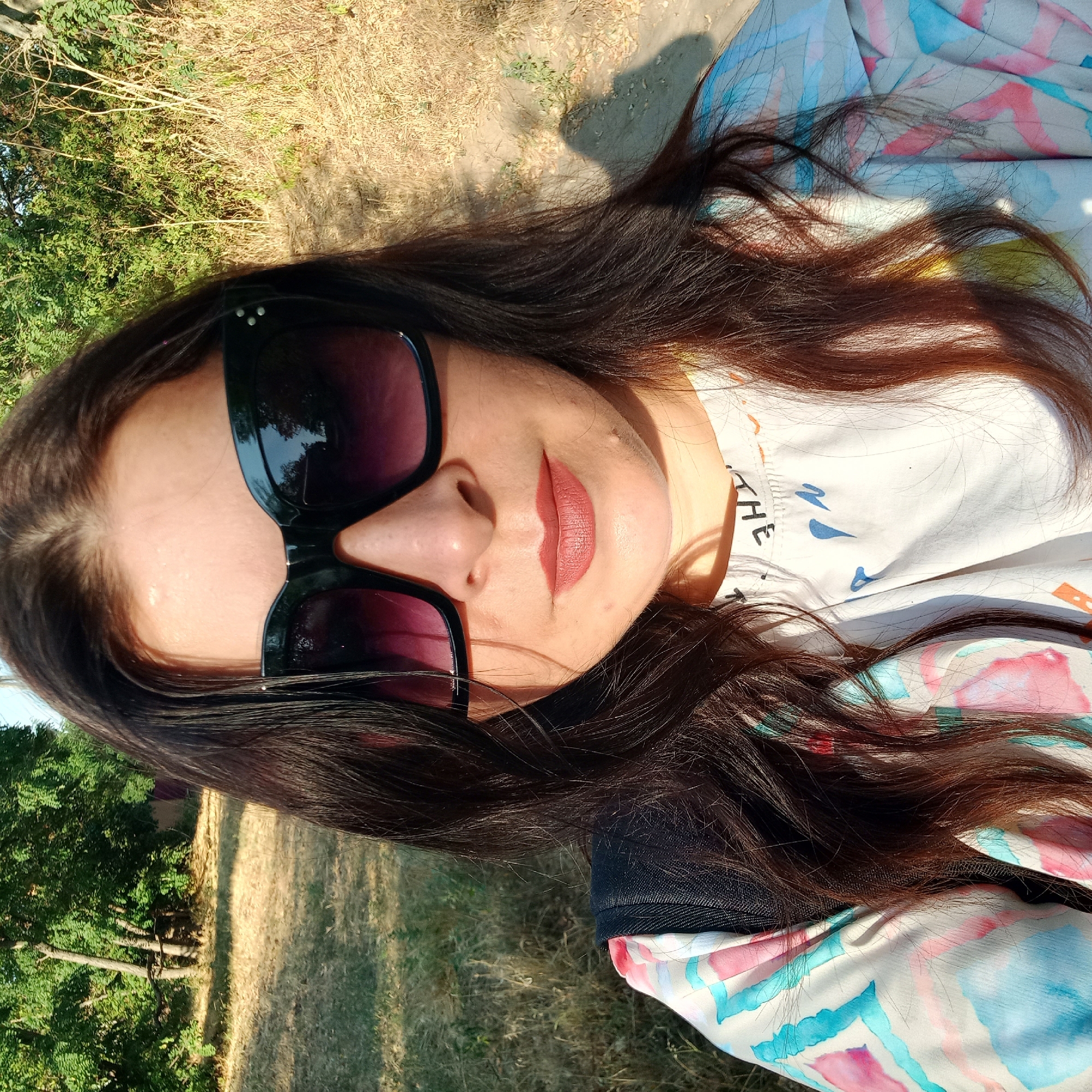 A woman with long, dark brown hair and large, dark sunglasses is pictured from the chest up, facing the camera. She is wearing a light-colored shirt with a watercolor-like pattern of pink, blue, and yellow shapes. The background suggests an outdoor setting, possibly a park or rural area, with dry grass and trees. The lighting indicates it is daytime, with sunlight casting shadows. There is no discernible indication of the specific location within Klyntsi, Ukraine, nor any context for the woman's presence or activity.