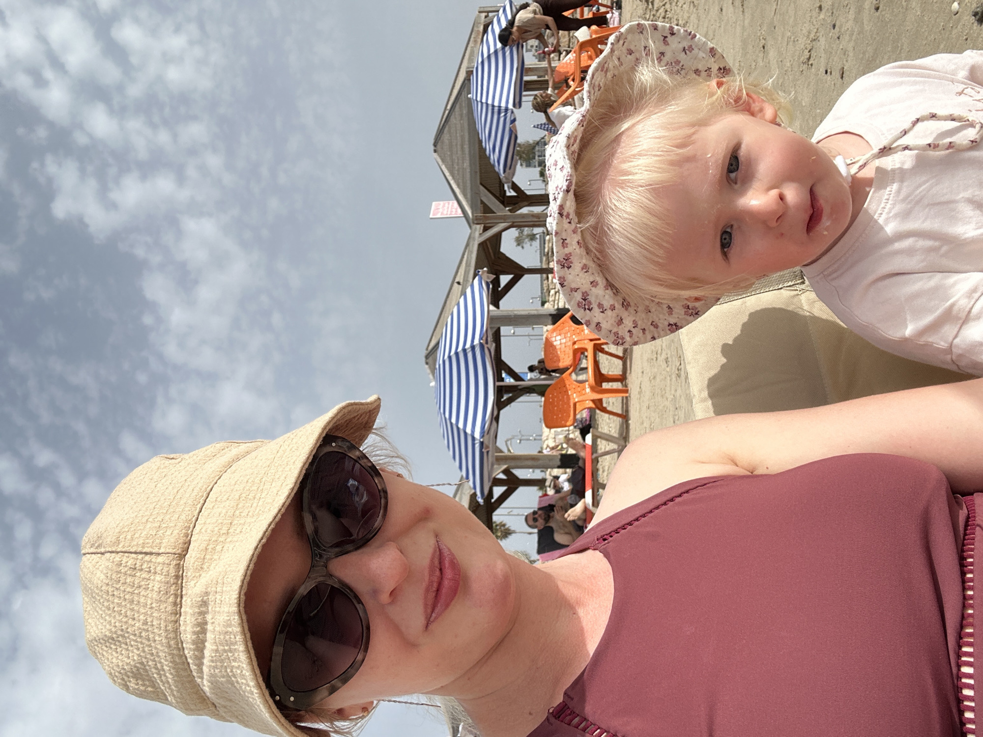 A selfie taken on a beach in Tel-Aviv, Israel shows a woman and a young child. The woman wears sunglasses and a tan-colored hat. She is wearing a maroon top with a ribbed edge. The child, who appears to be a toddler, is wearing a floral hat and has remnants of food on her face. They appear to be sitting. In the background are beach umbrellas with blue and white stripes, orange chairs, and other people. The sky is cloudy.