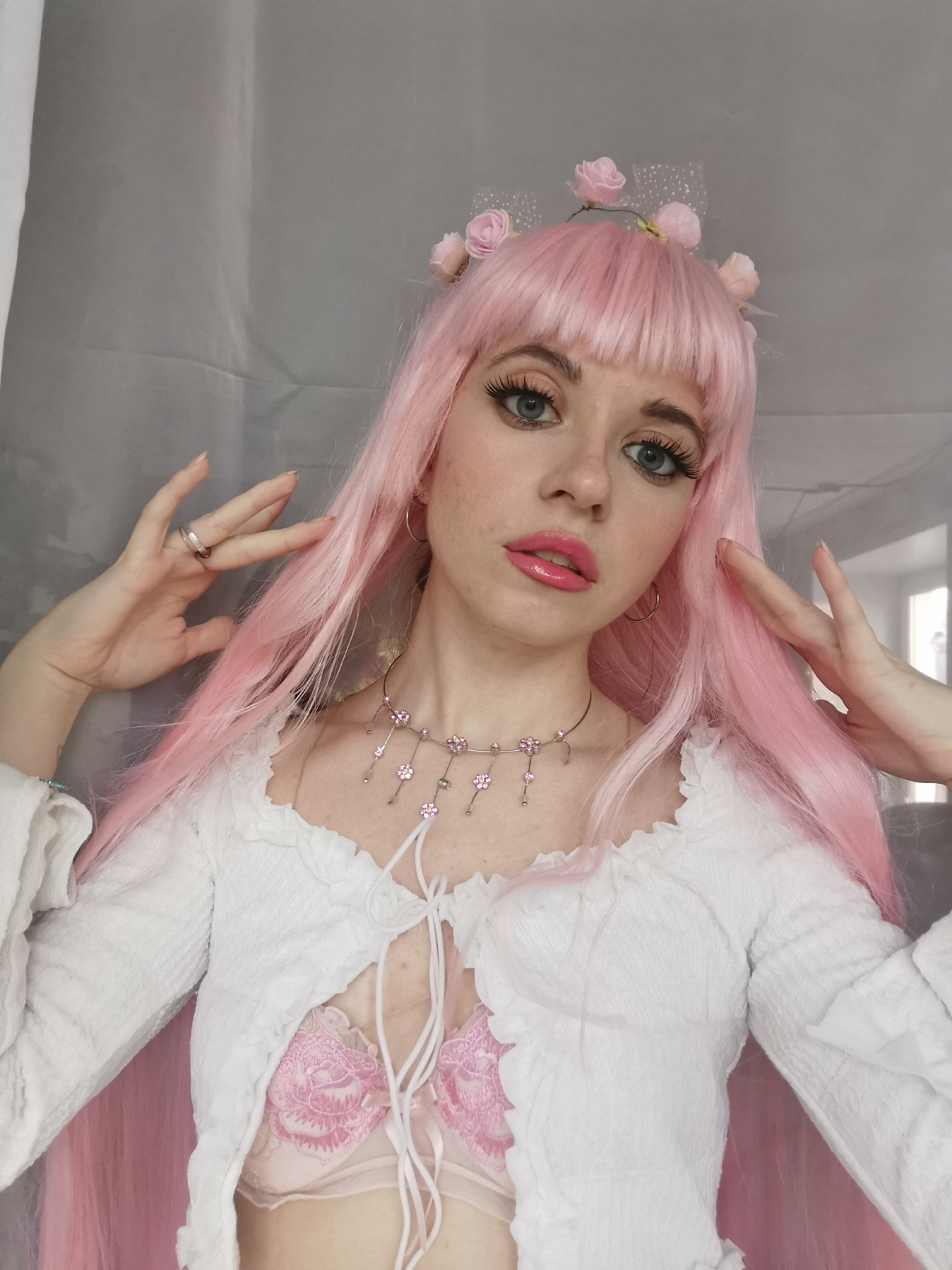 A young person, appearing to be a woman, is centered in a medium close-up, posing directly for the camera. She has fair skin, light blue-gray eyes, and wears a long, straight, bright pink wig with bangs. Her makeup includes prominent eyelashes, defined eyebrows, and shiny pink lipstick.
She is wearing a white, long-sleeved, cropped top with a textured or ruched fabric and ruffled trim around the neckline. The top features a tie-front closure, revealing a pink lace bra with a floral design underneath, as well as a glimpse of her midriff. On her head, she has a delicate headband adorned with small pink rose-like flowers and a sheer, sparkling mesh detail.
Her hands are raised and positioned symmetrically on either side of her face, with fingers slightly spread, featuring light-colored nail polish. A silver-toned ring is visible on the ring finger of her left hand. Around her neck, she wears a silver-toned choker-style necklace embellished with multiple small pink flower charms, some with dangling elements. Small hoop earrings are also visible in both ears.
The background is a simple, light gray or off-white fabric, possibly a curtain or sheet, suggesting an indoor setting. The lighting is soft and even, highlighting her features without harsh shadows, implying daylight or diffused artificial light. Her expression is neutral and confident, with a direct gaze. No visible text is present in the image.