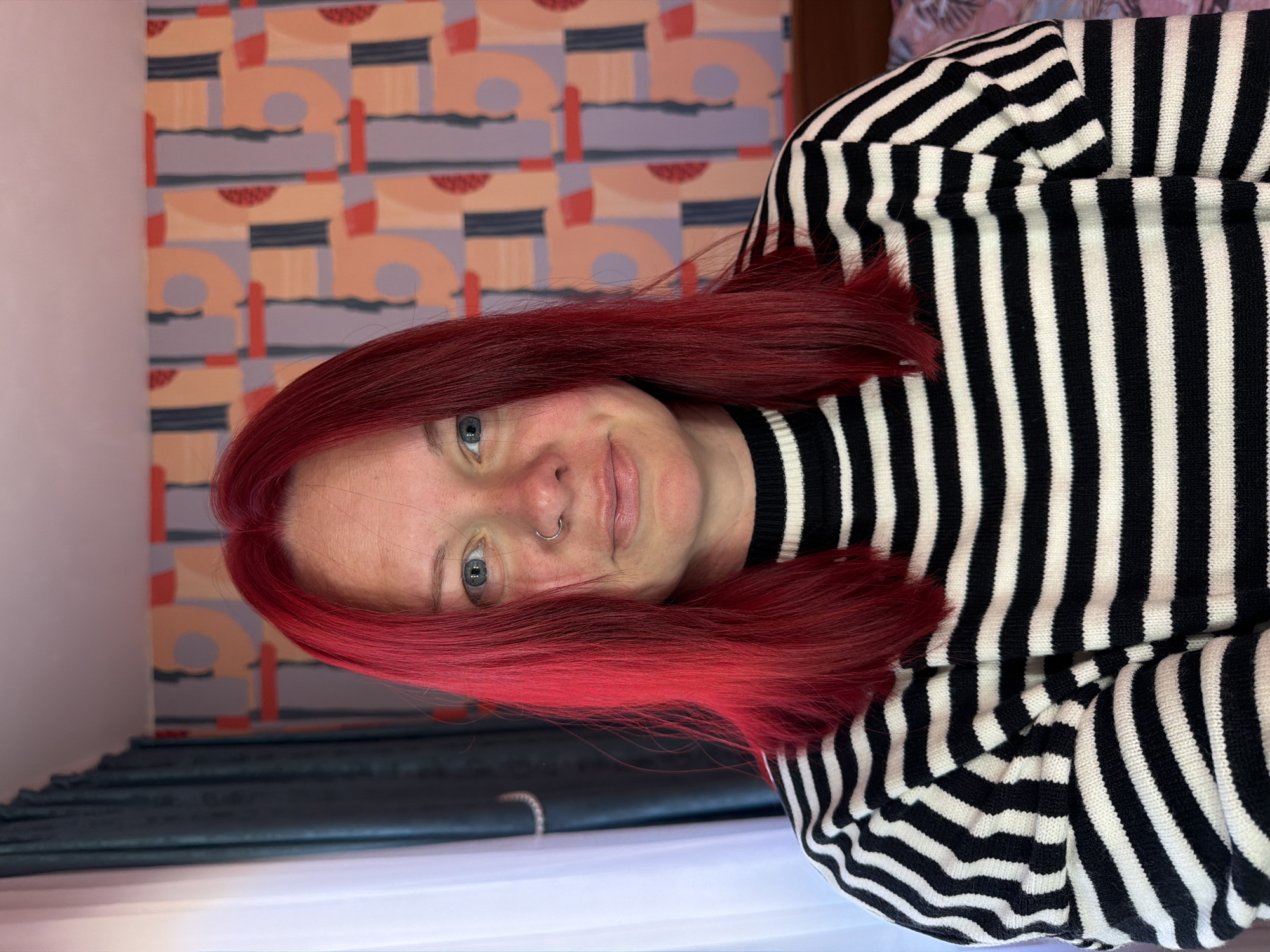 A woman with shoulder-length, bright red hair is the central figure in this photograph. She has fair skin, light-colored eyes, and a nose ring. Her expression is a slight, friendly smile. She is wearing a black and white striped sweater with a high neckline.
The background features a patterned wallpaper consisting of overlapping, abstract shapes in shades of gray, peach, and red on a muted base color. There is also a dark green curtain visible on the left side of the image, adding depth to the scene.
The photo appears to have been taken indoors, with the woman positioned in front of the wallpaper. The lighting suggests a natural source, potentially daylight.
There is no verifiable information in the image to definitively place the location in the city of Chaikovsky, Russia.