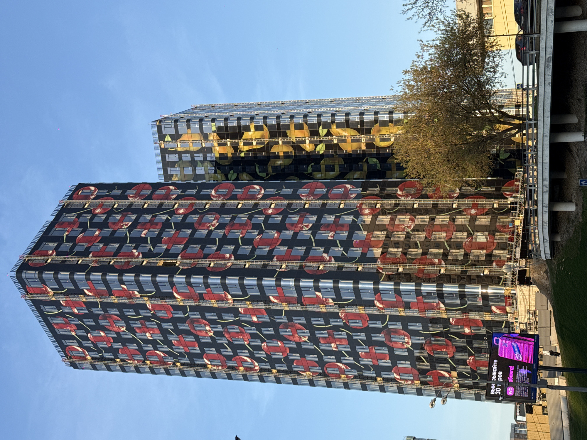 The image shows two tall buildings in Moscow, Russia. The closer building is decorated with a pattern of red cherries on a black background. The building behind it has a pattern of yellow lemons. A tree is visible between the buildings, partially obscuring the lemon building. To the right of the building a billboard is visible. The sky is blue and clear.