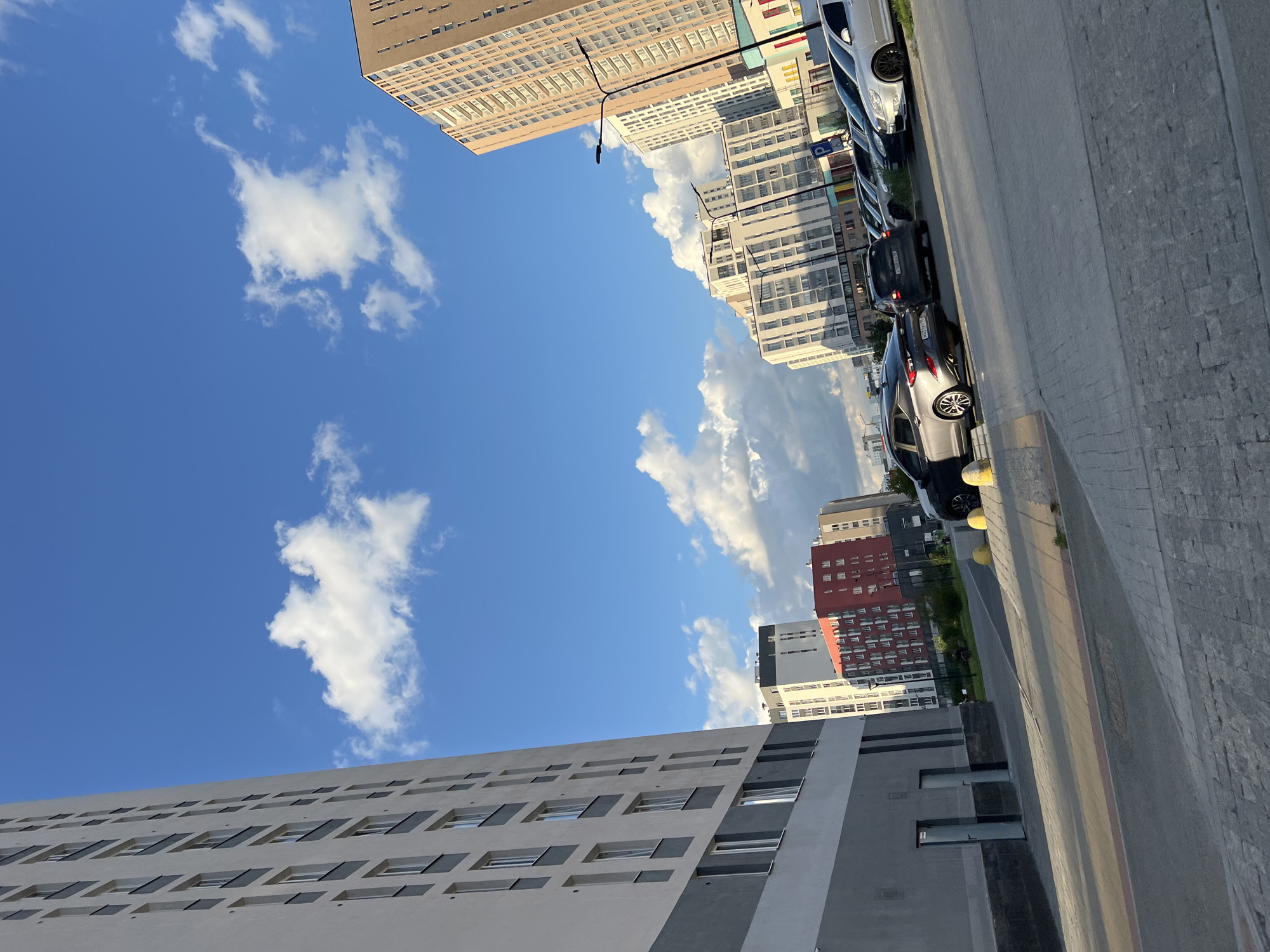 The image depicts an urban scene with a clear blue sky and scattered white clouds. Several modern apartment buildings of varying heights and architectural styles are visible. Some buildings are light-colored, while others feature red and grey facades. Many windows are visible on the buildings, suggesting residential units.
In the foreground, a paved walkway or road runs along the side of the buildings. Several cars are parked in a row along this path, including a silver sedan and a dark-colored SUV. Yellow bollards are placed along the edge of the walkway.
The perspective is from a low angle, looking upwards towards the sky and the buildings. The lighting suggests it is daytime, likely late afternoon or early evening, given the warm tones and shadows. The weather appears to be fair.
The scene is set in Yekaterinburg, Russia, as indicated by the provided location context. There is no visible text in the image that provides significant context or information. The overall impression is one of a modern residential urban environment.