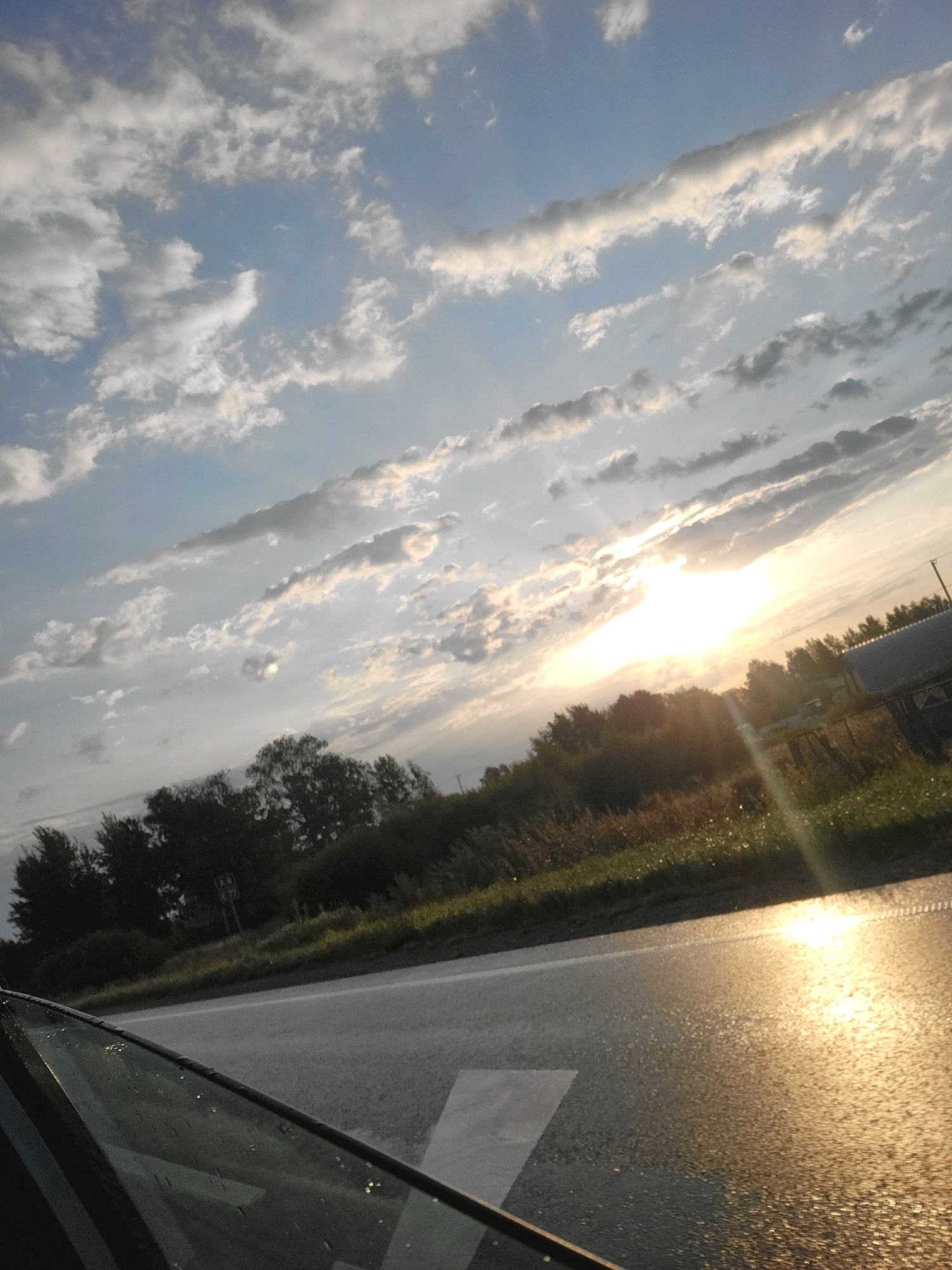 The image depicts a road with a car window in the foreground on the left. The road appears wet, reflecting the sunlight which is breaking through scattered clouds in a blue sky. Trees line the right side of the road, and beyond them, a structure is visible. The scene is indicative of a rural or semi-rural setting, possibly on a highway, with the sunlight suggesting either sunrise or sunset. The location is identified as Brankstūri, Latvia.