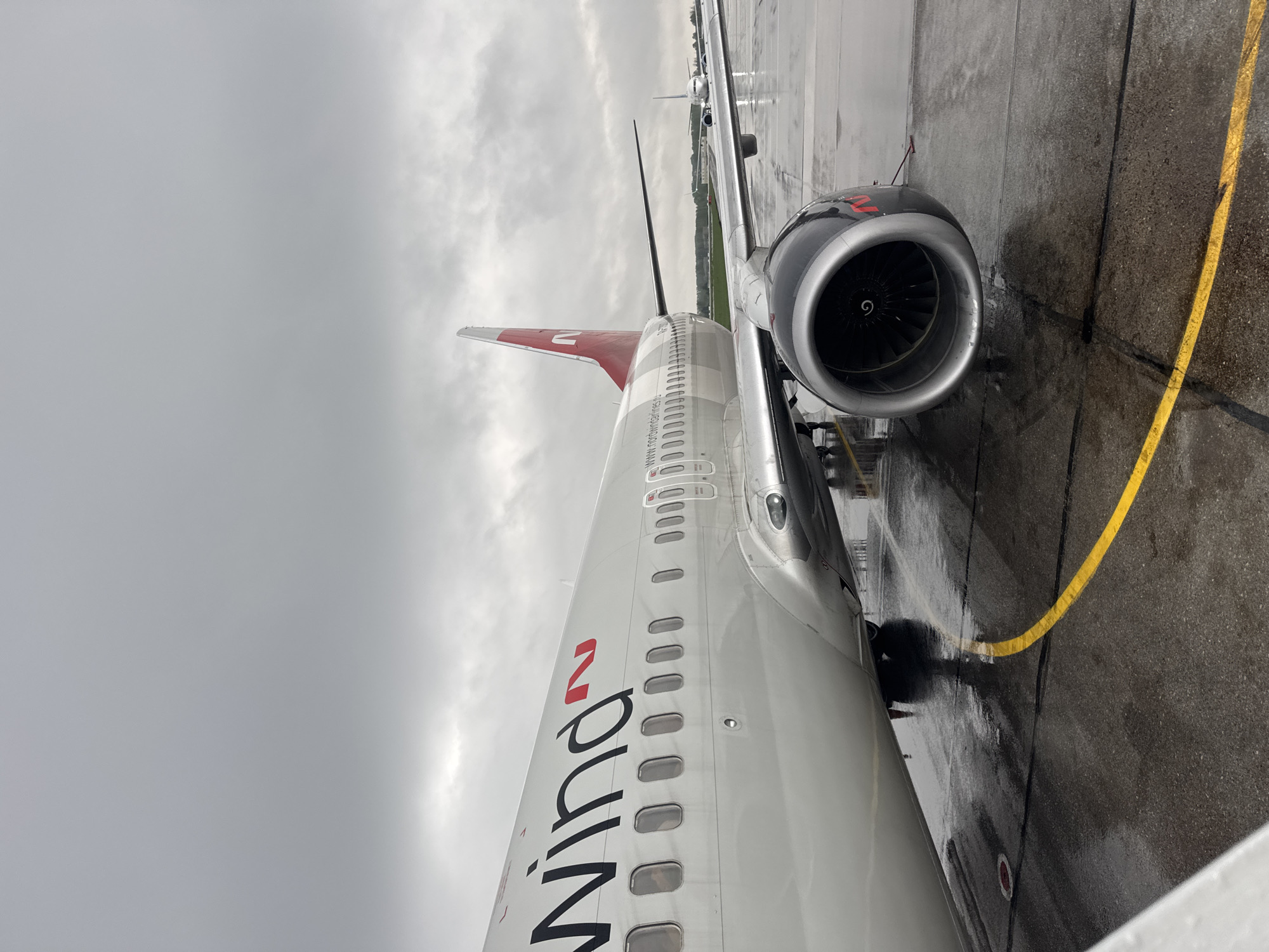 The image shows the exterior of a Nordwind Airlines airplane, possibly a Boeing 737, parked on a wet tarmac. The airplane is white with the airline's name in black and red lettering. Part of the tail fin, also in red and white, is visible. A jet engine is attached to the wing. A yellow line is painted on the tarmac. The sky is overcast, indicating a rainy or cloudy day. The airplane is potentially located at Bolshoye Savino Airport, situated near the city of Bolshoye Savino, Russia. There are no people visible.