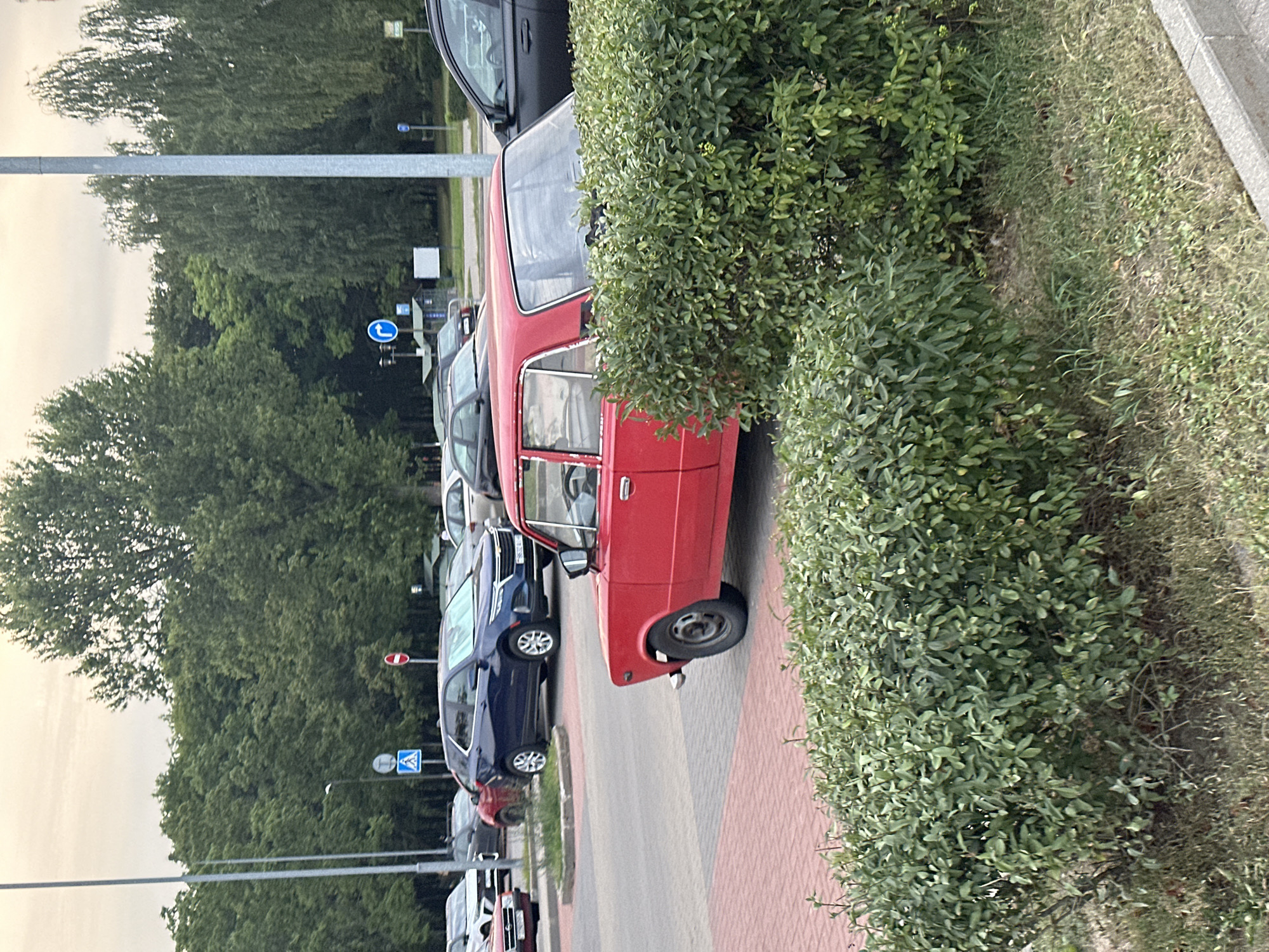 The image shows a parking lot with several cars and trees in the background. The sky is overcast. In the foreground, a red car is parked partially obscured by a green bush. The car appears to be a vintage model. Other cars, including a dark blue SUV and a black sedan, are visible in the background. There are also traffic signs visible, including a pedestrian crossing sign and a "no entry" sign. The ground is paved with bricks and asphalt. The overall scene suggests an outdoor parking area in a city, possibly a residential or commercial zone. It is likely located in Minsk, Belarus, given the presence of cars and signage that are common in the region.