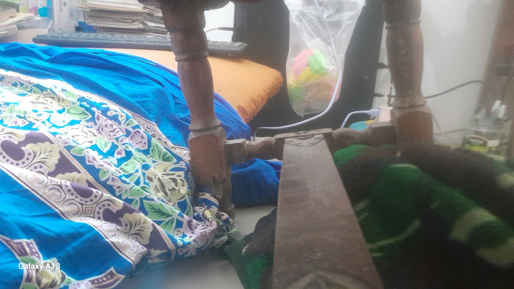 The image displays a cluttered indoor scene, likely a room, with various objects and fabrics. In the foreground, a blue fabric with a floral pattern in purple, green, and white is prominently visible, draped across a wooden structure. Behind this, a wooden chair leg is apparent, with another wooden beam running horizontally in front of it. To the left and behind, a bright orange surface is visible, possibly a cushion or mat, covered partially by folded blue fabric. Further back, a keyboard is seen on a flat surface, suggesting a desk or table. To the right, a clear plastic bag contains colorful spherical objects, possibly toys. Electrical wires and a power strip are also present, indicating electronic devices. The background is generally out of focus, but hints of shelves with stacked papers or books can be discerned. A white wall forms the backdrop on the right side. The overall impression is of a disorganized, domestic space. The location is identified as Hadipolo, Indonesia.