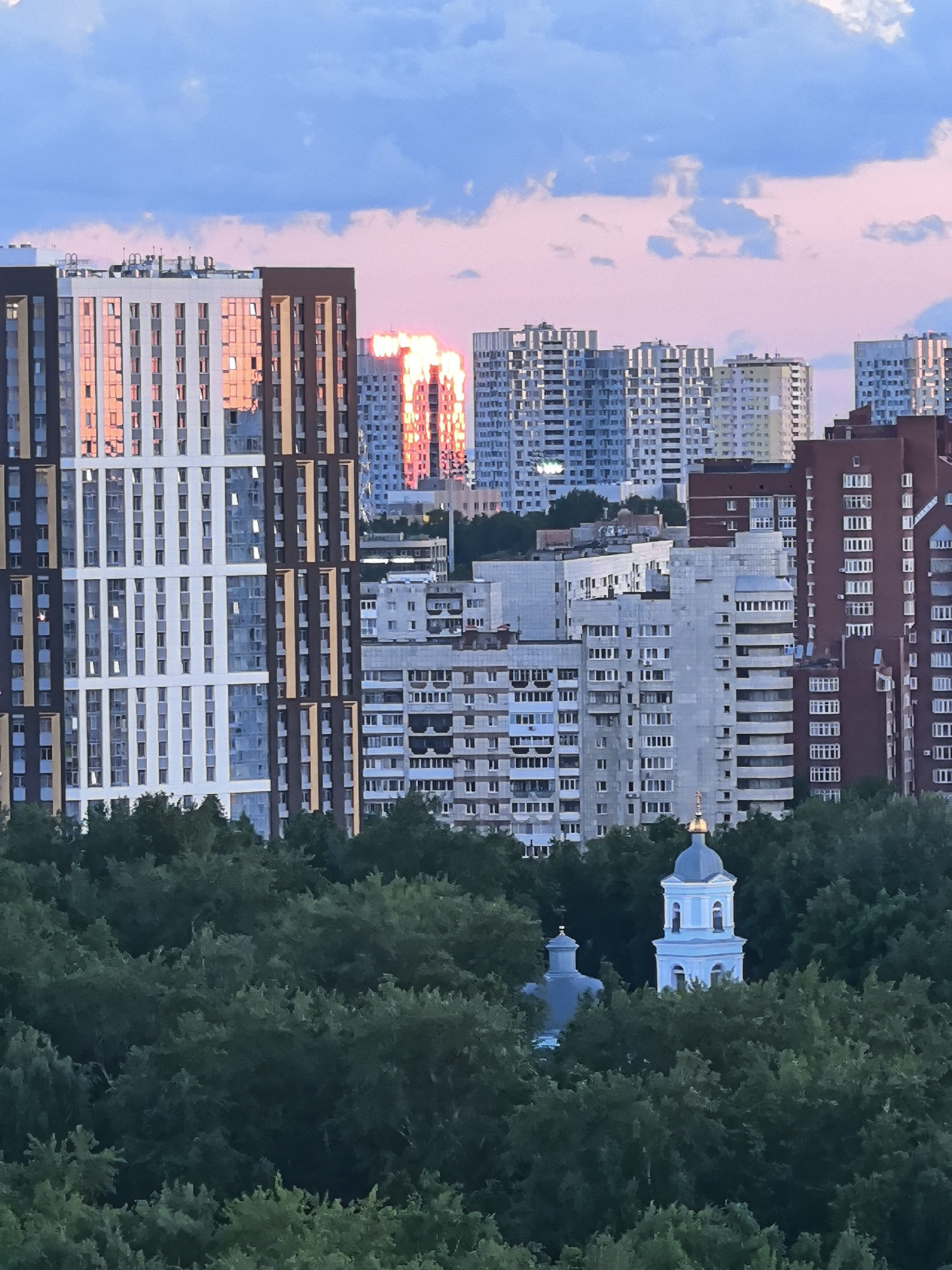 A wide-angle photograph captures a dense urban landscape in Perm, Russia, viewed from an elevated perspective, likely during either sunset or sunrise. The foreground and mid-ground are dominated by a lush, dark green canopy of deciduous trees, indicating a substantial forested area or parkland.
Nestled within and rising above the trees in the mid-ground is a light blue church steeple, featuring a golden onion dome and a cross, typical of Orthodox Christian architecture. A smaller, darker dome is partially visible to its left, suggesting a larger church complex.
Beyond the trees, a sprawling cityscape unfolds. A diverse collection of multi-story apartment buildings of varying architectural styles and ages populate the background. On the left, a prominent modern high-rise features sections of white and dark brown cladding, with many of its windows intensely reflecting a vivid orange-pink light from the low sun. Other tall buildings further back and to the right also exhibit these striking, fiery reflections on their facades. Interspersed among these newer structures are numerous older, more utilitarian gray and off-white residential blocks, consistent with Soviet-era design, creating a stark contrast in urban development.
The sky above presents a soft blend of blue and pink hues, with scattered white clouds, characteristic of either dawn or dusk. The lighting is diffused but the strong, warm reflections on the buildings indicate the sun is low on the horizon, casting a warm glow across parts of the city. No people or discernible activities are visible in the scene. There is no visible text. The overall impression is one of a vast urban environment harmoniously coexisting with natural elements, highlighted by the atmospheric lighting of a transitional time of day.
