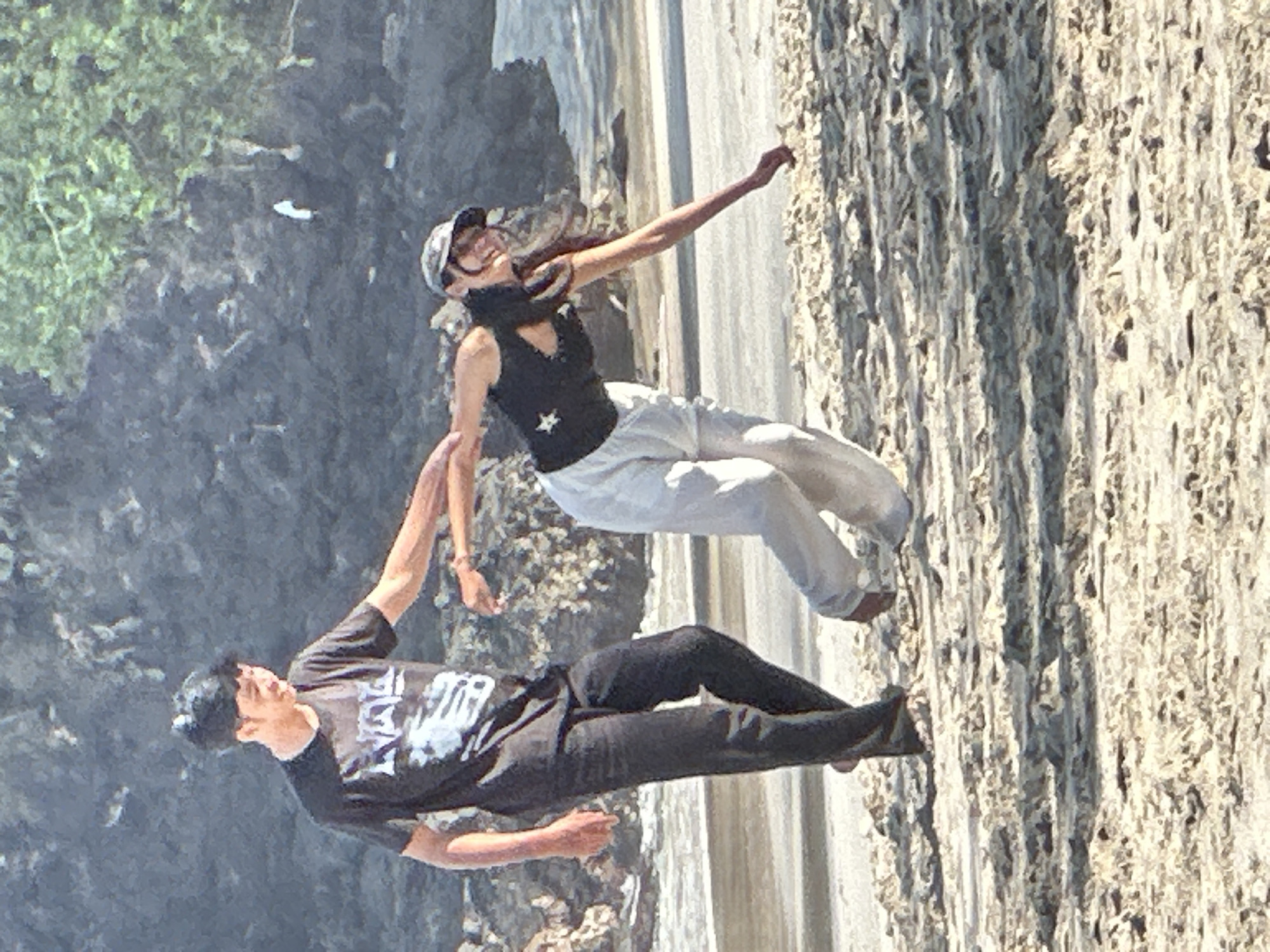 Two individuals are on a sandy beach, with a rocky, vegetated cliff face in the background and the ocean or water in the mid-ground. The person on the left, a male with dark hair, wears a black t-shirt with white lettering and dark pants. He is pulling the arm of the person on the right. The person on the right, a female wearing a black tank top, a baseball cap, and light-colored pants, is leaning away from the male and reaching towards the sand with her right hand. The scene appears to be outdoors, during daylight. The location is identified as Manggis, Indonesia.