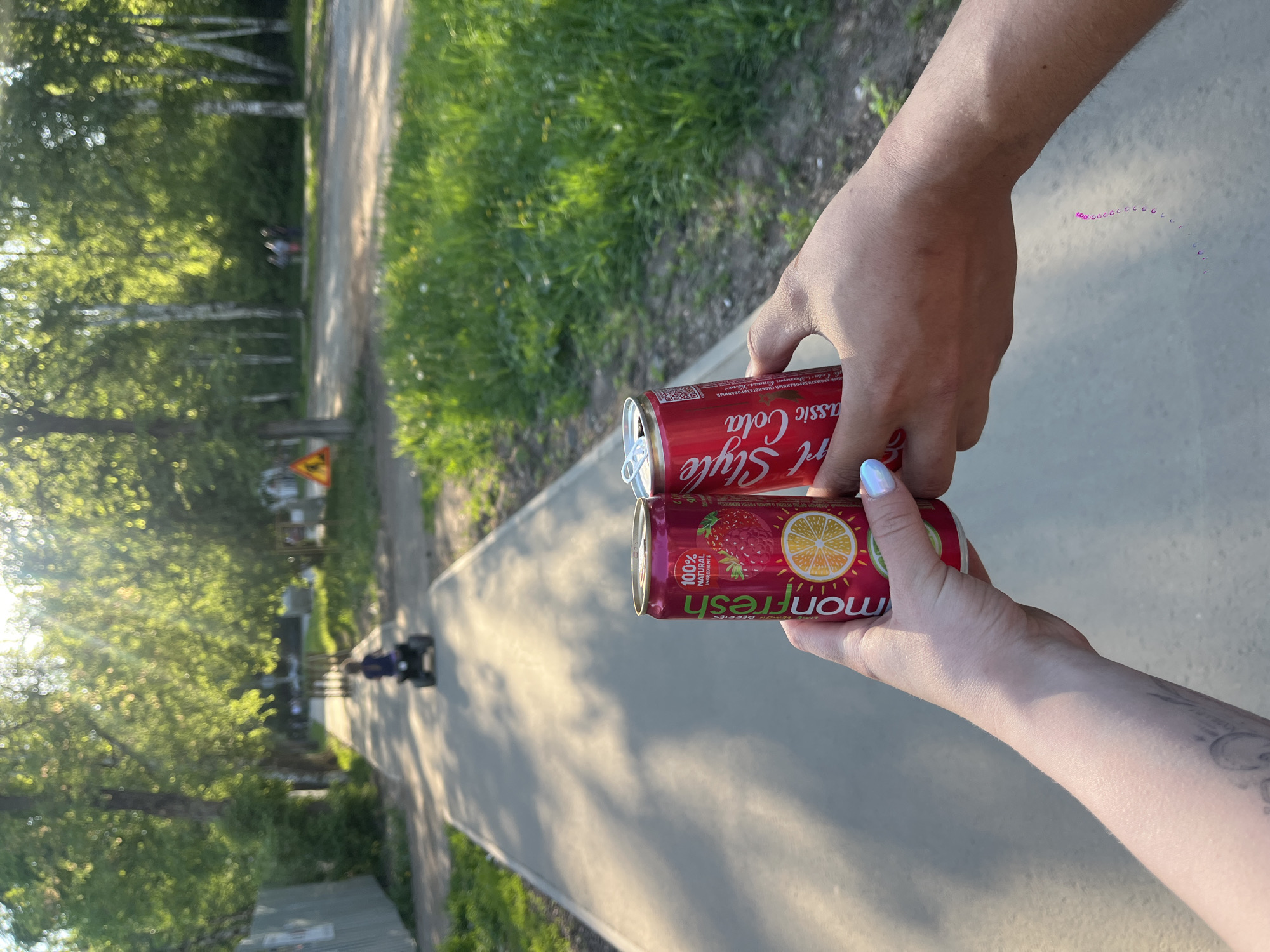 Two people are standing on a paved path in what appears to be a park or green area. Each person is holding a red can of a beverage. The person on the left is holding a can labeled "Art Style Classic Cola," and the person on the right is holding a can labeled "Monfresh" with an image of a strawberry and an orange slice. The path stretches into the background, where trees and green foliage are visible. In the background, a person is riding a scooter or a small vehicle down the path. Based on visual clues such as the style of the cans and vegetation, and the location of the image, it is likely located in Izhevsk, Russia.
