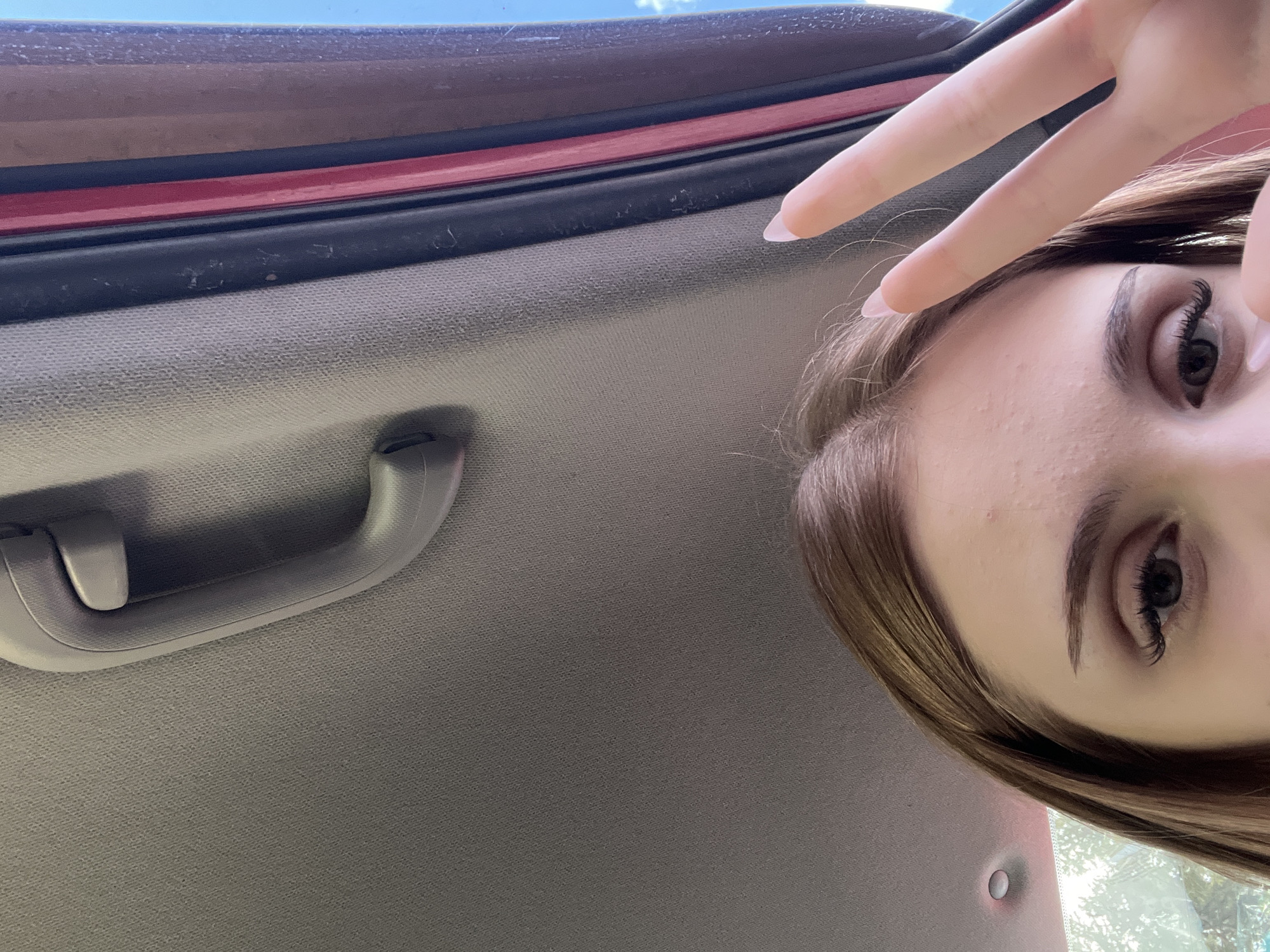 The image shows a close-up, low-angle shot of a young person's face and hand, taken from inside a vehicle, likely in the city of Tolbazy, Russia. The person has light brown hair and is looking towards the camera with one eye visible, the other partially obscured by a hand. Their right hand is raised in a peace sign gesture, with long, manicured fingernails. The interior of the vehicle's roof lining, a grey grab handle, and the edge of a red-painted door frame are visible in the background. The lighting suggests it is daytime, with sunlight filtering in from outside.