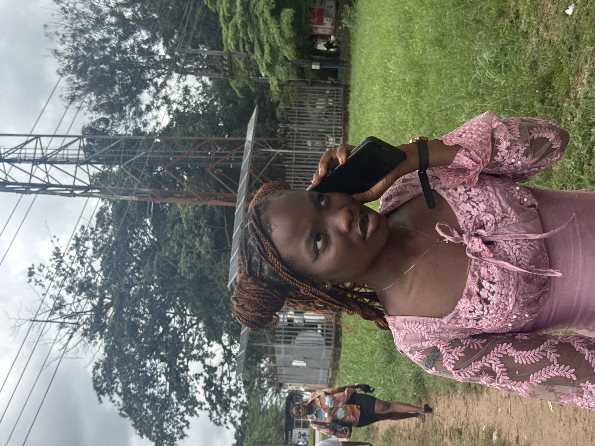 In Ihiagwa, Nigeria, a young Black woman with braided hair styled in two buns is captured mid-stride, holding a black smartphone to her right ear as if engaged in a conversation. She is wearing a pink, lace-detailed dress with a tie front. In the background, a dirt path curves to the left, where another Black woman in a patterned shirt and black skirt walks away from the camera, carrying a black handbag. A tall, metallic communication tower stands prominently on the right side of the frame. The environment is characterized by lush green grass, trees, and an overcast sky.