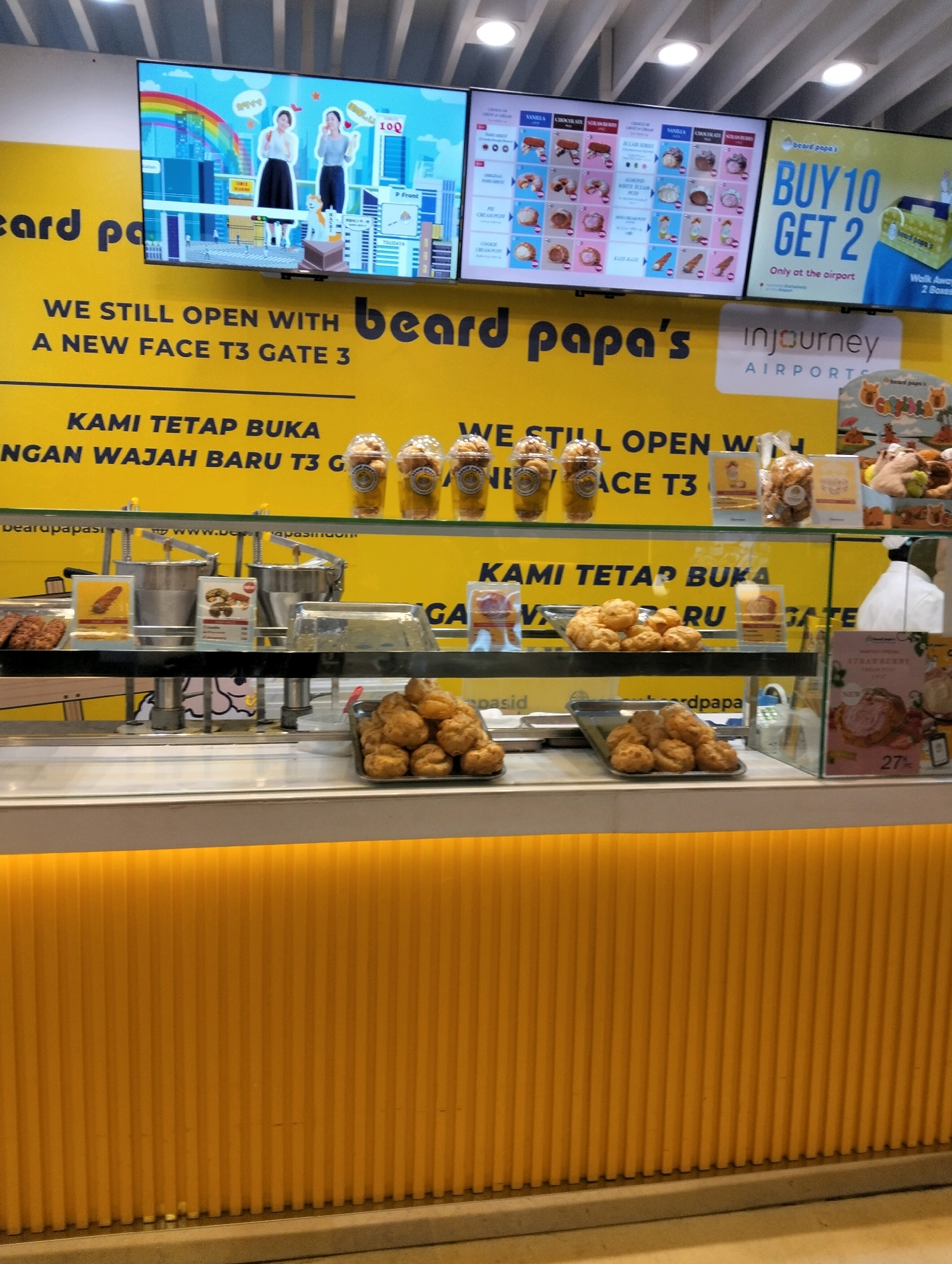 The image shows the interior of a Beard Papa's shop, likely located at an airport given the "T3 Gate 3" signage and the "inJourney AIRPORTS" branding. The shop features a bright yellow color scheme with vertical fluting on the lower half of the counter and walls. Two large screens display advertisements. The left screen shows animated graphics with two women and a rainbow, while the right screen displays product offerings and a "BUY 10 GET 2" promotion.
On display behind a glass counter are various cream puffs and baked goods. In the foreground, two trays hold piles of golden-brown cream puffs. To the left, a tray contains what appear to be fried pastries or fritters. Above the counter, several clear plastic cups filled with cream puffs are arranged on a shelf. A white-clad staff member is partially visible on the right side of the image, suggesting this is a functional retail space. The overall impression is a clean, modern, and brightly lit food establishment.