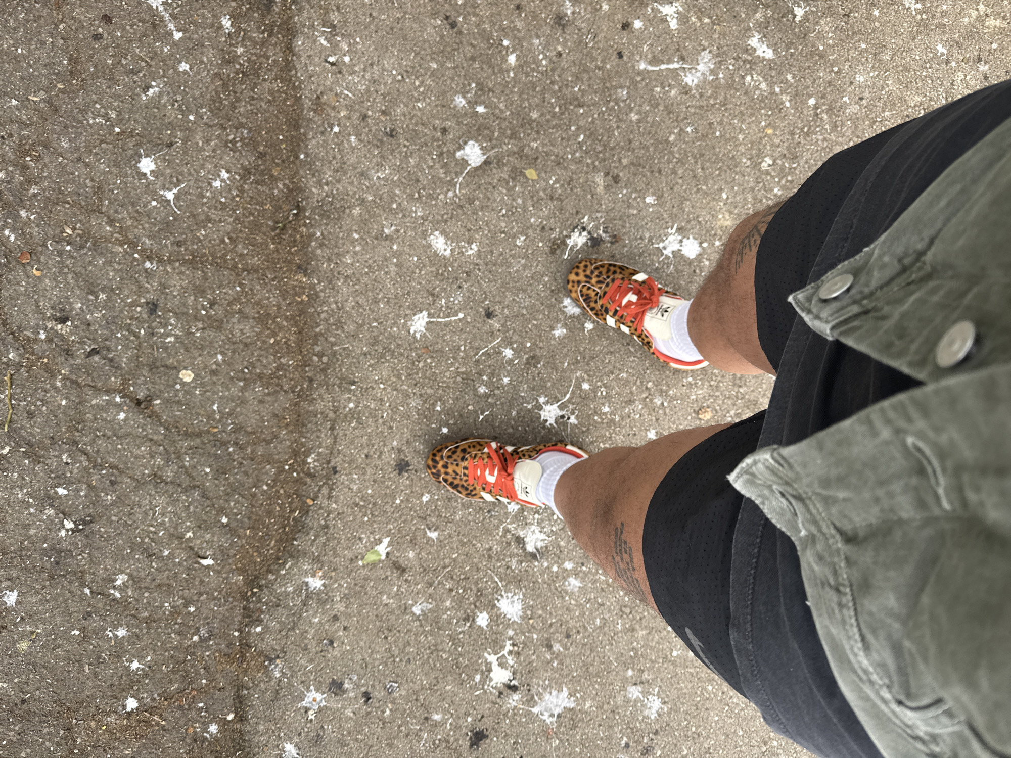 An overhead, high-angle shot captures a person's lower body and feet on a paved outdoor surface in Esch-sur-Alzette, Luxembourg. The individual is wearing black athletic shorts, white crew socks, and eye-catching leopard print sneakers with bright orange laces. A small "Adidas" logo is visible on the tongue of the right shoe. Part of a green or khaki utility jacket is visible on the right side of the frame, draped over the person's hip. A tattoo is discernible on the left leg. The ground, likely asphalt or concrete, is a mottled grey and is extensively covered with numerous white splatters, consistent with bird droppings. The bright, even lighting suggests it is daytime. The image depicts a moment of walking or standing, with attention drawn to the footwear and the distinctive, speckled ground.