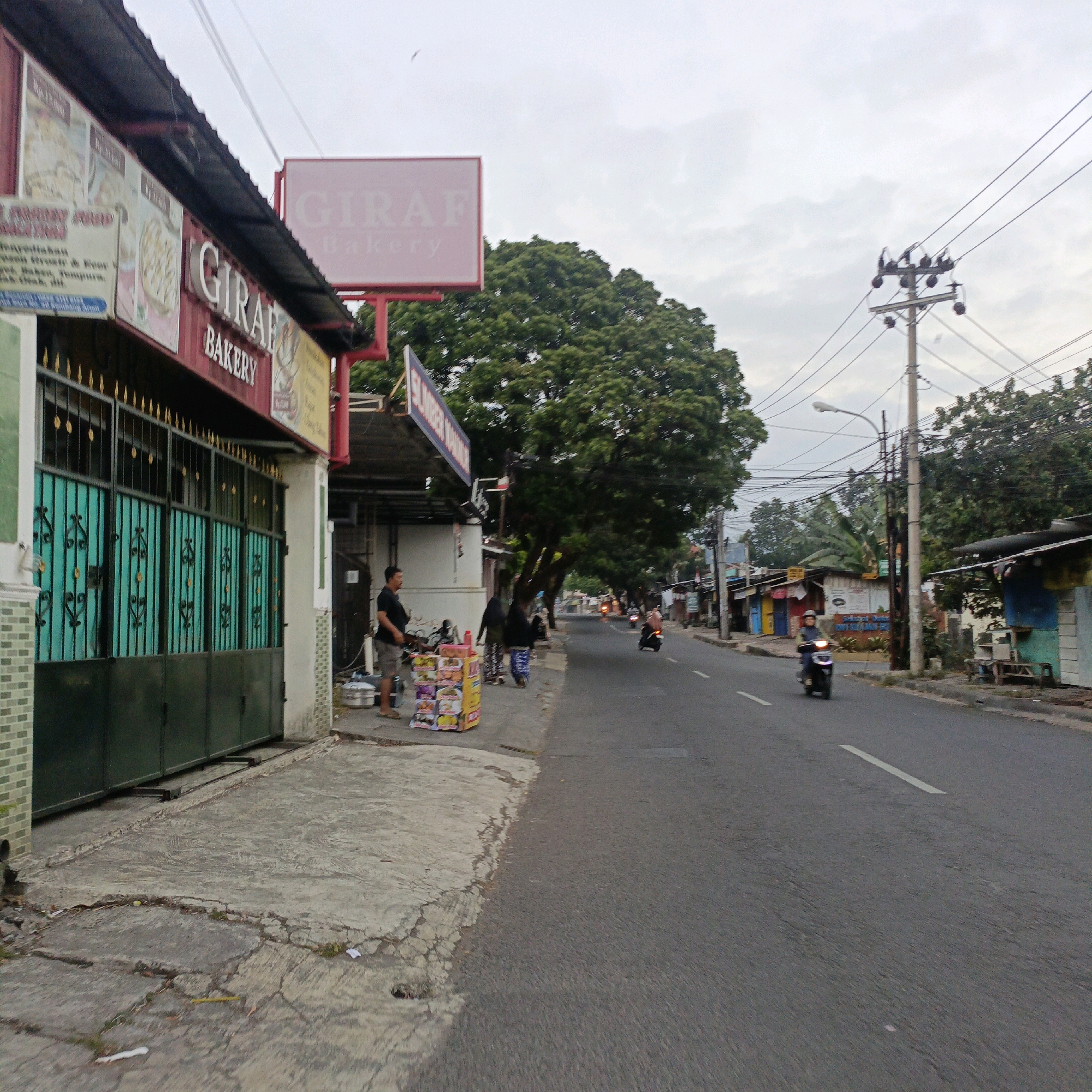 The image captures a street scene in Salatiga, Indonesia, likely in the morning or late afternoon given the soft lighting and overcast sky. On the left, a building with a red awning and the prominent sign "GIRAF BAKERY" is visible. The bakery has a green metal gate with decorative elements. A man in a black t-shirt and shorts stands to the right of the bakery's entrance, near a display of colorful boxes that appear to be food items. Further down the sidewalk, a street vendor with a patterned skirt and headscarf walks away from the bakery, and another person is seen walking in the distance.
The street itself is paved and has lane markings. Several motorcycles are on the road; one is in the foreground on the right, ridden by a person wearing a helmet. In the background, a few smaller buildings and stalls line the opposite side of the street, with trees providing shade over parts of the sidewalk and road. A utility pole with multiple wires is a prominent feature in the middle ground. The overall impression is a typical urban street in Indonesia with commercial activity and daily life unfolding.