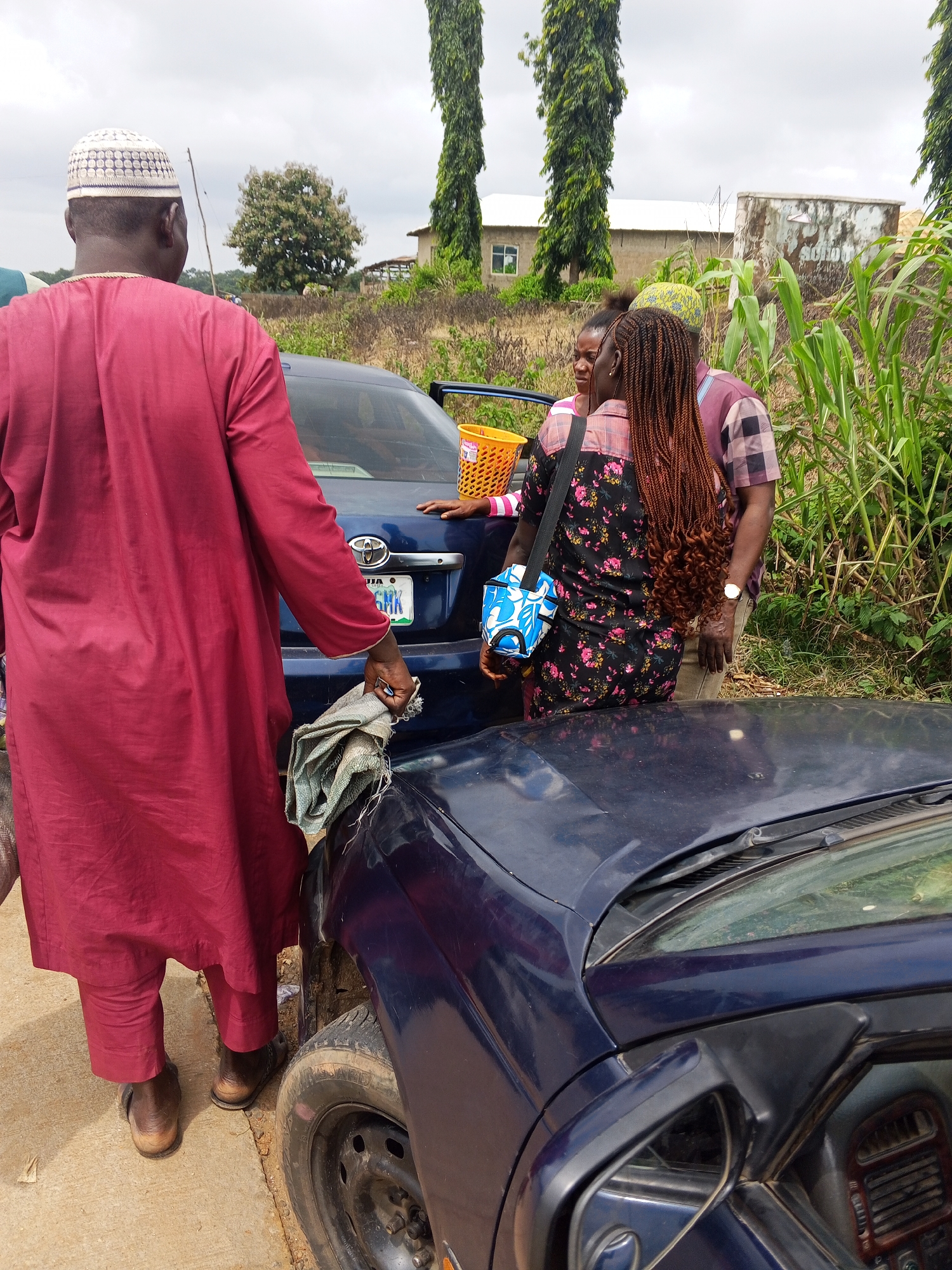 In the city of Oduape, Nigeria, under an overcast sky, three individuals are gathered near a dark blue sedan. A man in a maroon robe and white cap stands facing away from the camera, holding a folded grey cloth. To his right, two women are positioned near the open driver's side door of the car. One woman, with long braided hair, wears a floral dress and has a blue and white bag slung over her shoulder. The other woman, partially visible, is behind her. A yellow basket sits on the car's trunk. The car's license plate is partially obscured. The scene unfolds amidst lush green vegetation, including tall trees and corn stalks, with distant buildings and a concrete structure visible in the background. The ground is a mixture of concrete and dirt.