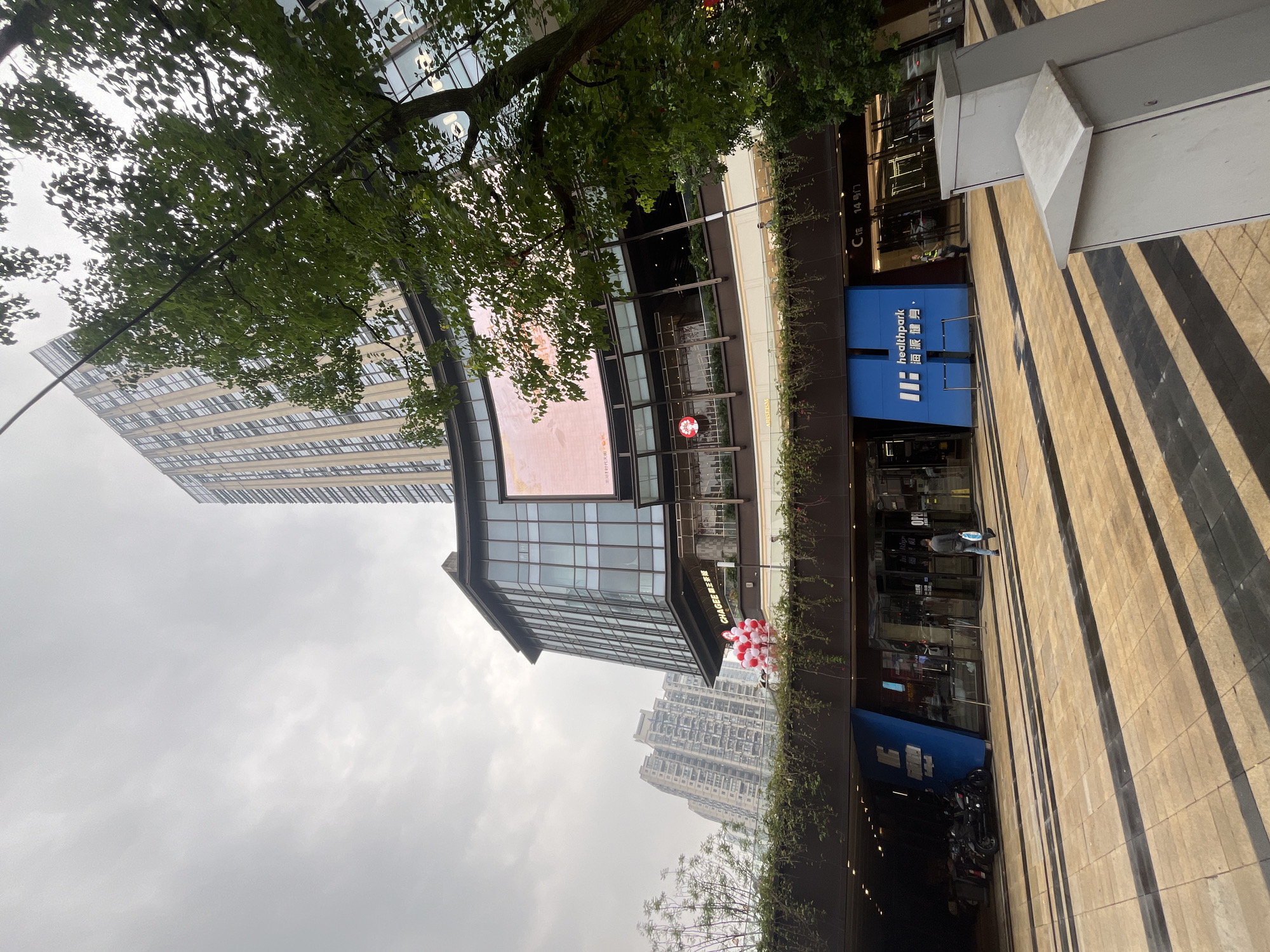 The image shows a street scene in Yuzhong District, China. A modern, multi-story building with a large glass facade dominates the background. In front of it, a lower-level commercial area has a bright blue entrance with "healthpark" signage. A man in a dark jacket and jeans is walking away from the camera, carrying a light blue bag. To the left of the blue entrance, several motorcycles are parked under an overhang. A tree with lush green leaves partially obscures the view of the building on the right side of the frame. The ground is paved with light-colored tiles, with darker rectangular patterns. The sky is overcast and gray.