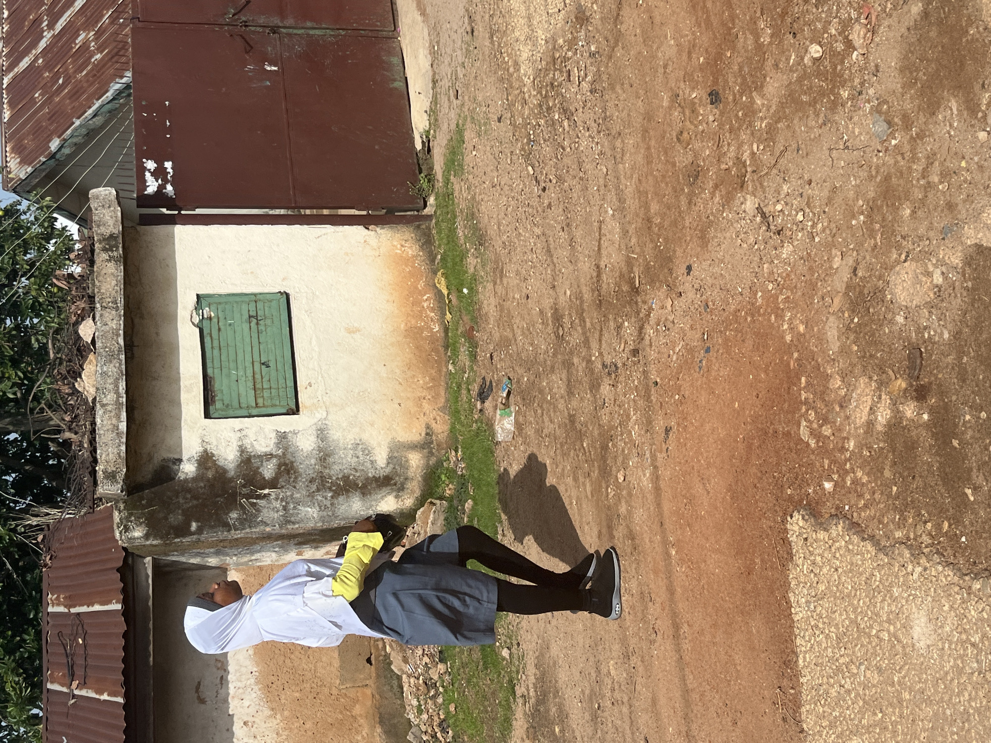 In the city of Jos, Nigeria, a young person wearing a white hijab, a blue skirt, and black leggings stands on a dirt road in front of a simple building with a rusty corrugated metal roof. The building has a white-washed exterior, partially covered in moss and grime, with a small, green, louvered window. A large, dark red metal gate is visible to the right of the building. The ground is a mix of dry earth, gravel, and scattered debris, including a plastic bottle. The person is wearing yellow gloves and appears to be holding a dark object. The scene is illuminated by natural daylight under a partially cloudy sky.