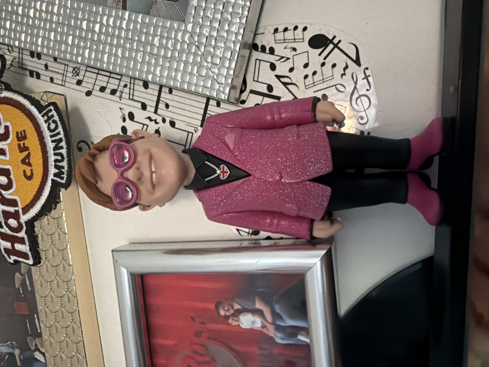 The image shows a small Elton John figurine wearing a pink glittery suit, black pants, and pink boots, placed on a white surface near a picture frame containing a photograph of a couple. Behind the figurine are a "Hard Rock Cafe Munich" patch and decorative items with musical notes. The city of Pétange, Luxembourg, is not evident in the provided image, and there is no other information that suggests its presence.