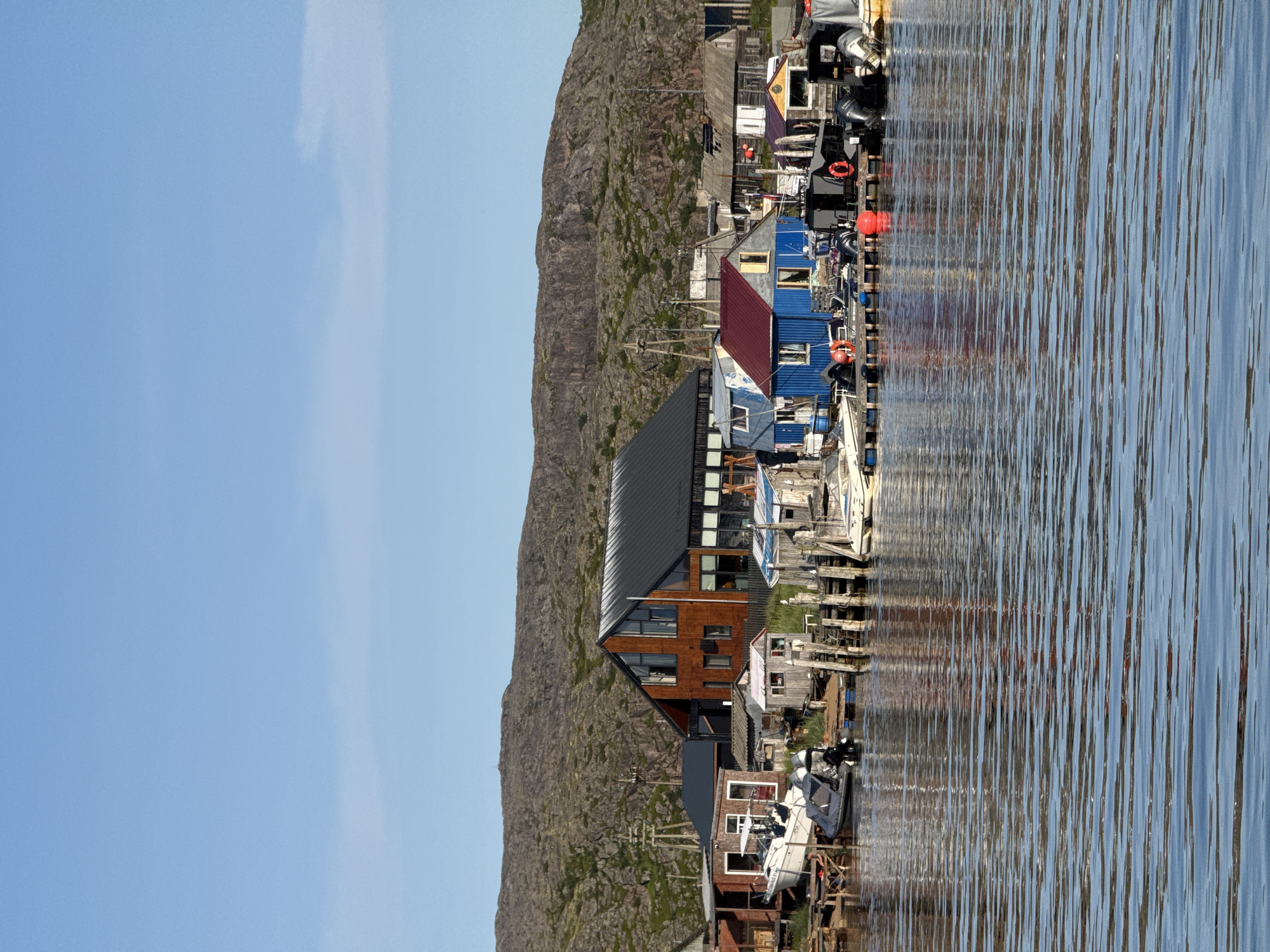 A waterfront scene in Teriberka, Russia, features a collection of houses, boats, and a rocky hillside under a clear blue sky. The houses vary in style, with one prominent modern building with a wooden facade and large windows. Other structures include smaller, older buildings with corrugated metal roofs and wooden exteriors. Several boats are docked, ranging from small motorboats to larger vessels. A wooden pier with pilings runs along the waterfront, supporting the docked boats and providing access to the buildings. The water in the foreground is rippled, reflecting the colors of the sky and the structures on the shore. The background is dominated by a rugged, rocky hillside with sparse vegetation. The overall impression is one of a coastal village or settlement.