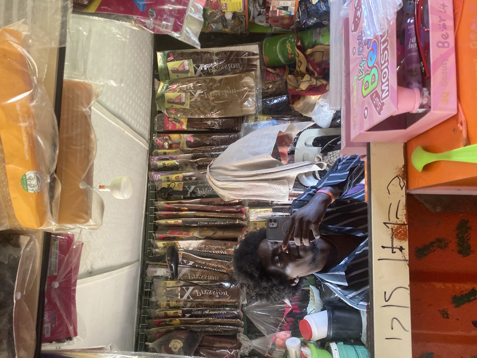 The image shows a man with dark skin and an afro-style haircut, wearing a striped shirt and holding a smartphone near his face. He appears to be taking a selfie in front of a shelf stocked with hair extensions of various shades. There is also a pink box labeled "MOIST!" and "Beauty Bear Life" visible. The products and their packaging suggest a beauty supply store. The date "12/5 + 1 = 2" is written on a surface near the items. Based on the presence of hair extensions and beauty products, it's likely that the photo was taken in a beauty supply store, possibly located in Lagos, Nigeria, based on the likely context and visual clues of product styles and types commonly found there.