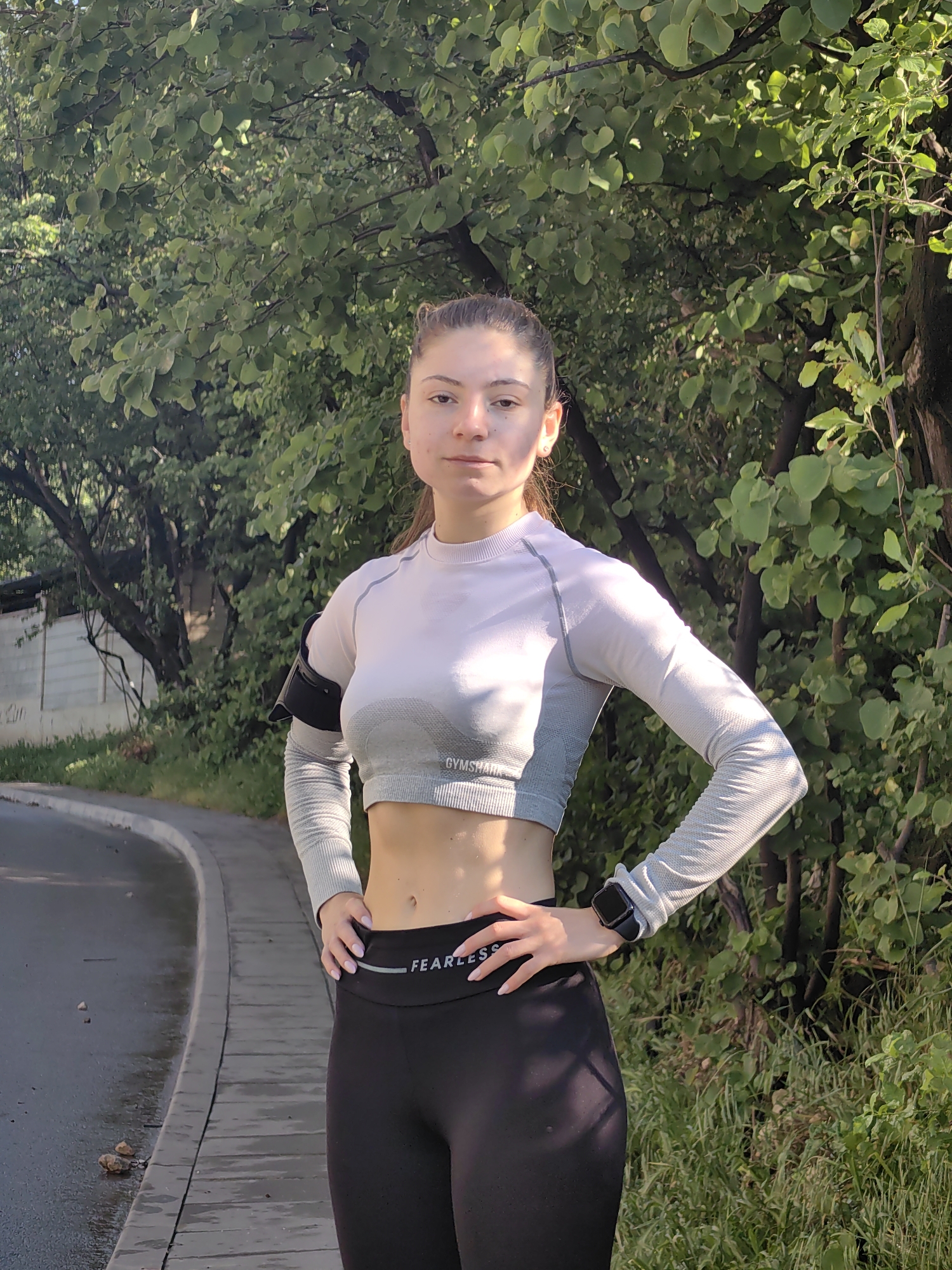 A young woman stands outdoors in what appears to be a park or green space. She is wearing workout attire, consisting of black leggings with the word "FEARLESS" on the waistband, and a cropped long-sleeve shirt. She has an armband on her left arm and a smartwatch on her right wrist. Her hands are placed on her hips.
The background features trees and vegetation, suggesting a park or natural environment, and part of a curb and street is visible. It's difficult to ascertain the exact location within Varna, Bulgaria, without more specific visual clues related to landmarks or characteristic features of the city.