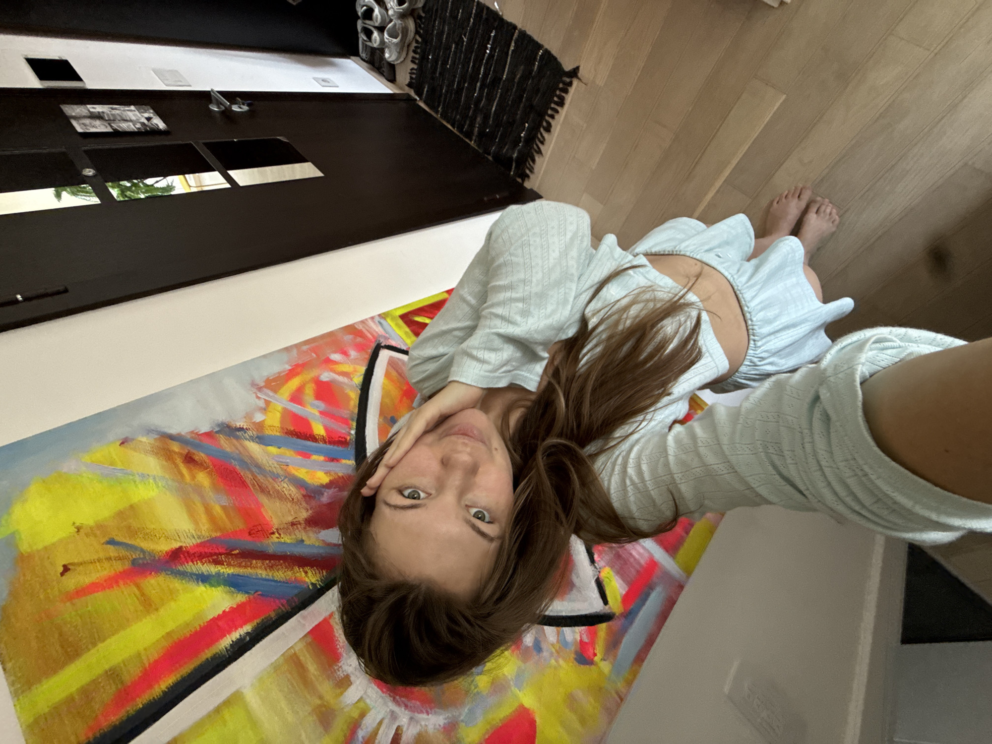 A woman is lying on the floor in what appears to be an apartment. The floor is made of wood. She is lying on a painted canvas, wearing a light blue outfit. Behind her is a door and a rug. No location information available to suggest this is in Vozdovac Urban Municipality, Serbia.