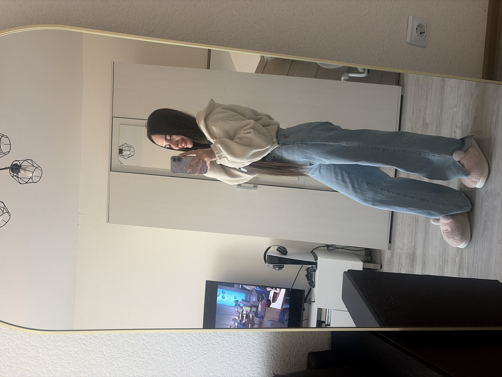 A young woman is taking a selfie in a full-length mirror. She is wearing a cream-colored, fluffy hooded sweatshirt, light blue jeans, and pink furry slippers. She is holding a smartphone in her left hand. The mirror is framed in gold. The reflection in the mirror shows a room with light-colored walls and a wooden floor. A light fixture with a geometric design is visible on the left side of the mirror. Behind the woman, there is a white wardrobe, a television showing an animated scene, a white shelf with a PlayStation 5 console, and a dark brown cabinet. An electrical outlet is visible on the wall. The location of the image cannot be determined.