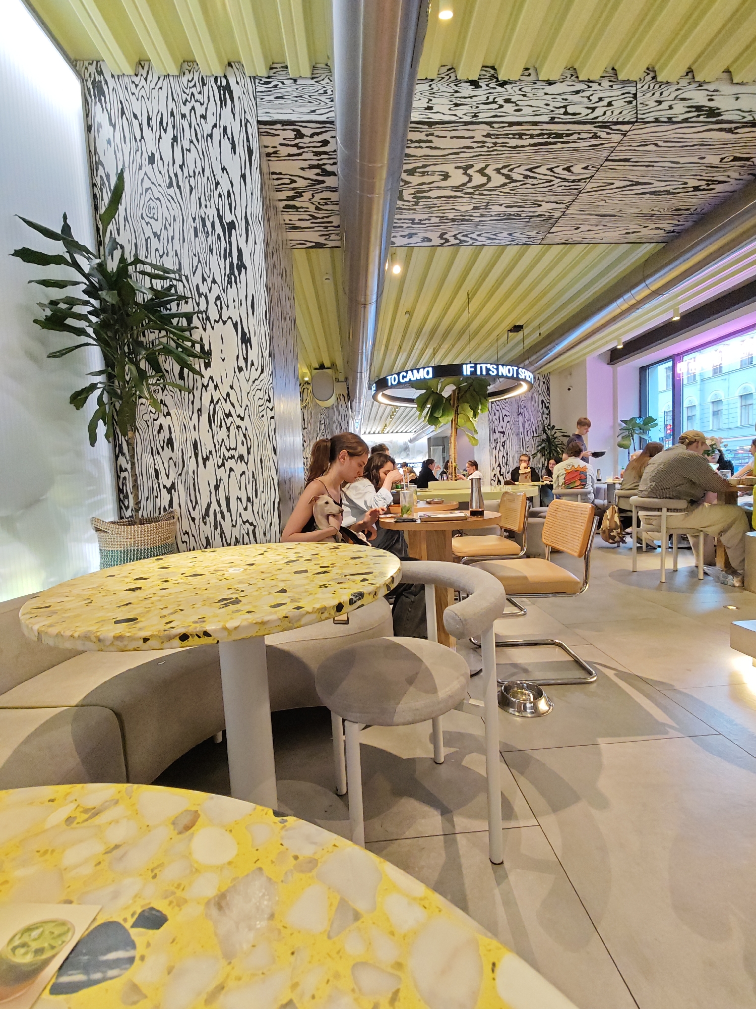 The image depicts the interior of a modern, brightly lit cafe in Saint Petersburg, Russia. The cafe features distinctive decor with a yellow corrugated ceiling and walls adorned with a black and white wood grain pattern. Tables are round and feature a terrazzo-style top with yellow, white, and black flecks. Chairs are upholstered in light grey fabric.
In the foreground, a large round table with a yellow terrazzo top is prominent, with a grey upholstered chair to its right. Further back, another similar table is visible with a woman sitting at it, holding a light-colored dog. Several other patrons are seated at tables throughout the cafe, engaged in conversation or eating. One server is seen in the middle distance carrying a tray. The cafe has large windows on the right side, offering a glimpse of the street outside with buildings visible. A circular neon sign is suspended from the ceiling, displaying text. The overall atmosphere appears casual and contemporary.