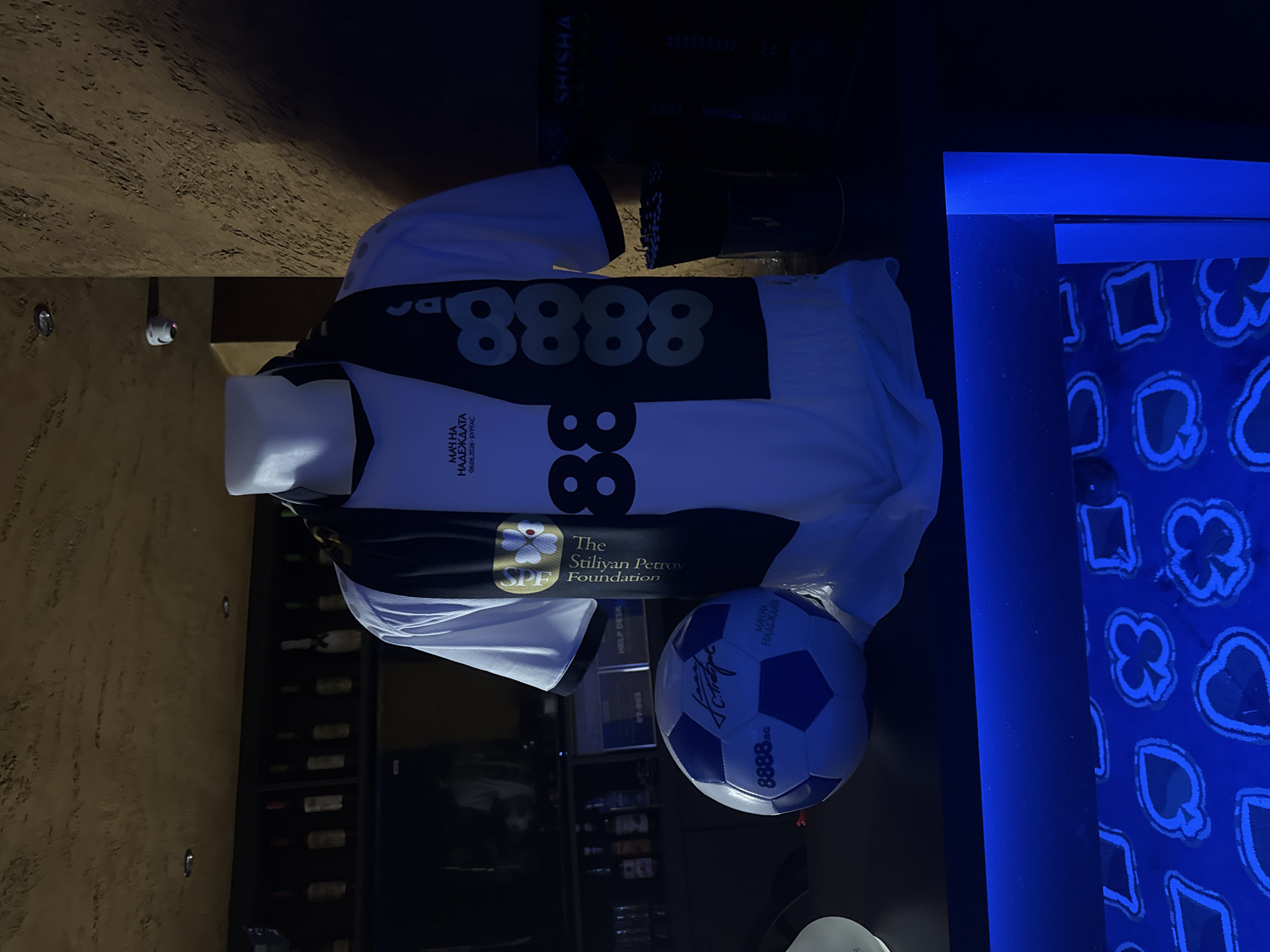 The image shows a football shirt and a soccer ball, likely displayed in an interior setting, possibly a bar or a sports-related establishment. The shirt is predominantly white with black accents and features the number "8888" across the chest. Below the number is the inscription "МАЧ НА НАДЕЖДАТА 06.06.2026 - БУРГАС", indicating a charity football match called "Match of Hope" taking place on June 6, 2026, in Burgas, Bulgaria. The "Stiliyan Petrov Foundation" logo is visible, suggesting an affiliation with the event. The soccer ball is blue and white, also branded with "8888.bg", and carries a signature. In the background, there are bottles in a rack, a "SHISHA" sign, and a blue patterned surface featuring playing card suits.