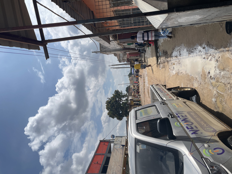 An outdoor scene in Luanda, Angola, captures a bustling unpaved street or alleyway on a partly cloudy day. The ground is muddy and filled with numerous puddles, suggesting recent rainfall. In the foreground on the right, a silver utility truck is prominent, parked or driving along the muddy path. The truck displays "NEONE" in purple lettering, along with the phone number "(+244)935 137 585" and colorful graphic logos. Other visible text on or near the truck includes "MUPS", "MELHOR", "DE CASA", and "GRA".
Overhead, a metal canopy structure with a corrugated roof extends into the frame, with power lines or communication cables visible stretching across the street. In the middle distance, several people are walking along the road; one individual wears a striped shirt, and another a yellow shirt. Buildings of various architectural styles, some featuring red or orange facades, line both sides of the street. A large green tree stands further down the road, and the street appears to continue into the distance with more buildings and activity. The bright sky with puffy white clouds indicates it is daytime.