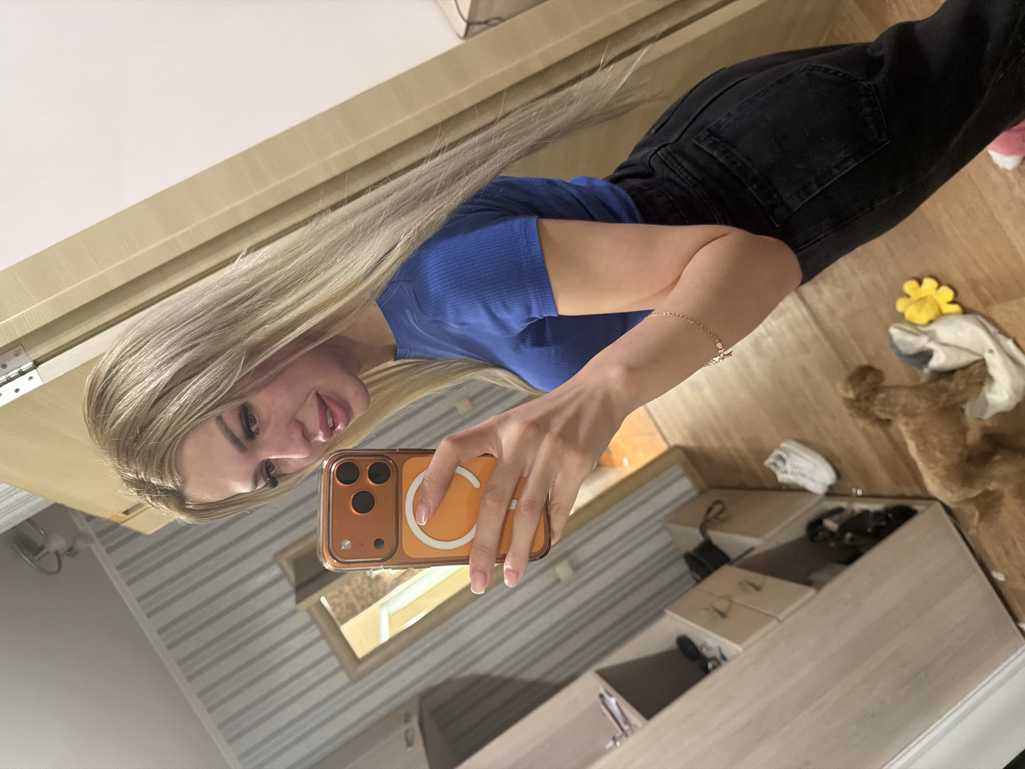 A young woman with long blonde hair, wearing a blue ribbed t-shirt and black jeans, is taking a mirror selfie in what appears to be a residential hallway or dressing room. She is holding an orange iPhone in her right hand. A small, fluffy dog and a yellow flower are visible on the floor. The environment includes wooden furniture, a mirror reflecting a doorway, and striped wallpaper. The flooring is wood. Based on the style of the interior and common residential architecture, the location is likely in Kyiv, Ukraine.