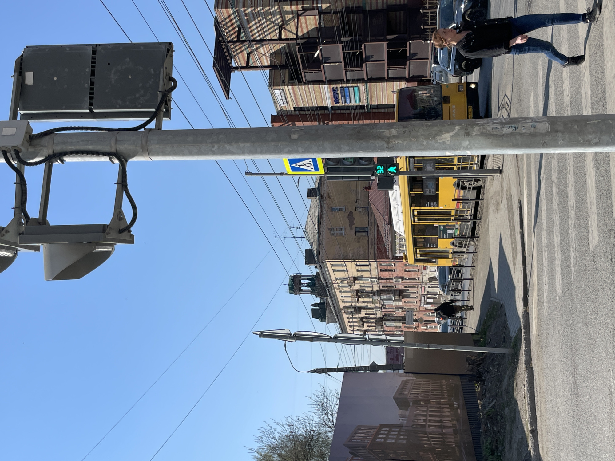 The image depicts a street scene in Izhevsk, Russia, showing pedestrian and vehicle traffic. A woman wearing jeans and a black jacket is crossing the street on a zebra crossing. A yellow public transport vehicle is positioned on the right side of the road. A street sign with a pedestrian crossing symbol is mounted on a grey pole with traffic lights. In the background are multi-story buildings. Overhead power lines stretch across the clear blue sky. A small patch of trees is visible in the bottom-left corner.