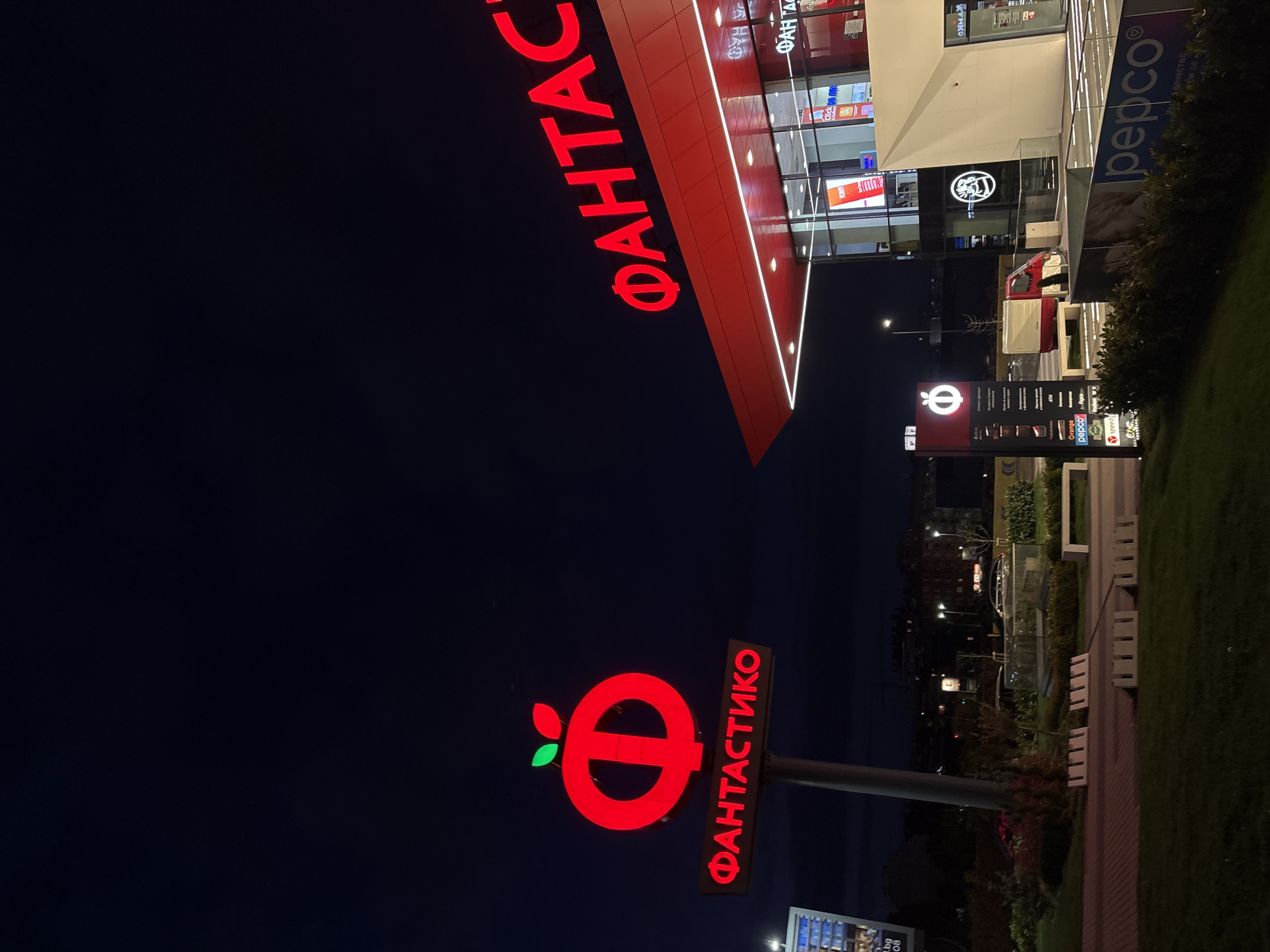 The media file captures a nighttime exterior view of a commercial building, likely a shopping center, in Sofia, Bulgaria. The scene is dominated by bright, illuminated signage against a dark sky.
The main subject is a modern, multi-story building featuring large red letters spelling "ФАНТАСТИКО" (Fantastico), a well-known Bulgarian supermarket chain, running along its roofline. The building also displays the logo for "pepco" on one facade and offers services like "Click & Collect." Its glass windows reflect the surrounding lights.
In the foreground, to the left of the main building, stands a tall, vertical illuminated sign. This sign prominently features Fantastico's distinctive red apple-like logo, followed by "ФАНТАСТИКО" written vertically. Below this, a directory lists other businesses or services, including "Orange," "pepco," and "speedy," among others like "KORFF," "OHMAX," and an "ATM."
The ground level around the building is landscaped with green grass, shrubs, and trees. White, minimalist benches are visible along a paved walkway. A light-colored truck or delivery van is parked near the building. Streetlights illuminate parts of the ground and the distant background, where other structures can be faintly seen against the dark sky. The overall impression is a bustling commercial area after dusk, with artificial lights providing the primary illumination.