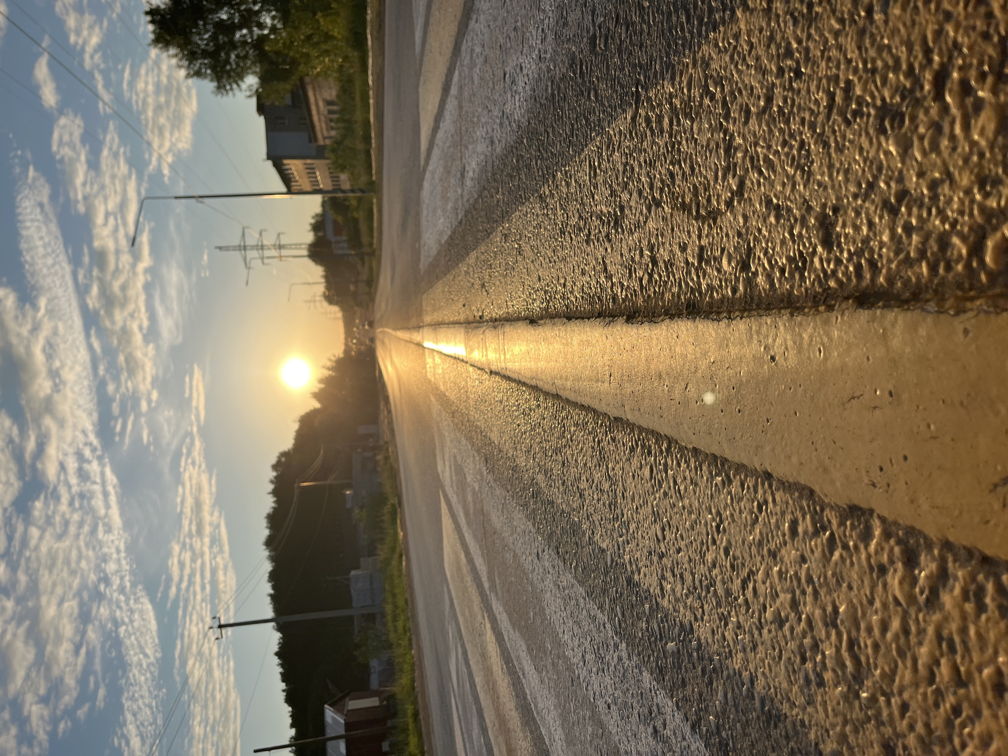 This image depicts a low-angle view of an asphalt road at either sunrise or sunset, illuminated by a low-lying sun positioned toward the left of the frame. The exact location within the city of Chaikovsky, Russia, cannot be determined from the visible elements.
In the immediate foreground, the textured surface of the asphalt road is prominent, featuring a distinct double yellow line running centrally into the distance, heavily reflecting the golden sunlight. To the left of this line, white pedestrian crossing stripes are visible, also reflecting light. The road surface appears to have a coarse, granular texture.
The road recedes into the midground, with the sun's reflection creating a bright path along its surface. Utility poles with overhead power lines are visible along both sides of the road, extending towards the horizon. Distant, indistinct vehicles are faintly visible on the road surface. A tall power pylon is discernible on the right side of the midground.
In the background, a dense line of trees forms a dark silhouette against the horizon on the left. On the right, several low-rise buildings, appearing to be industrial or older institutional structures, are visible amongst some foliage. The sky above is partly cloudy with scattered white cumulus clouds against a pale blue backdrop.
No individuals or specific human actions are present within the frame. The scene is characterized by the strong, warm, directional lighting from the sun, which is a bright orb near the horizon.