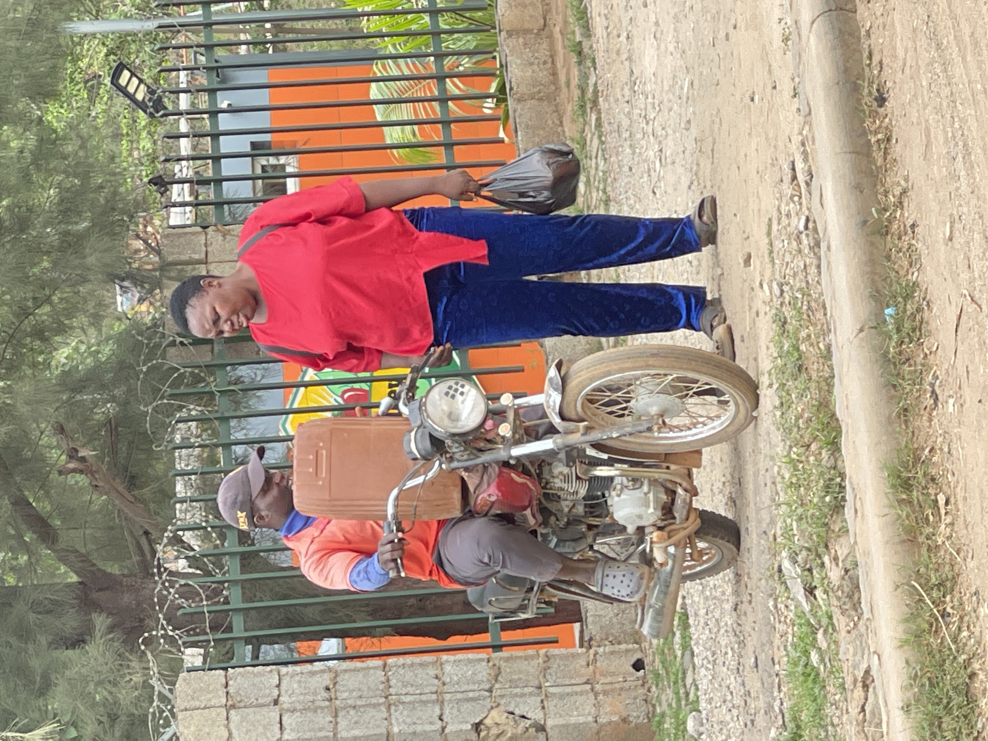 This outdoor image features two people and a motorcycle on what appears to be an unpaved road or dirt path.
A woman, standing on the right, wears a red long-sleeved top and dark blue patterned trousers. She holds a black plastic bag in her right hand and looks downwards. To her left, a man is seated on an older model motorcycle, holding the handlebars. He wears an orange long-sleeved shirt with blue undersleeves, dark trousers, a grey baseball cap, and grey Crocs. A light brown, rectangular container or package is secured in front of him on the motorcycle.
The motorcycle has spoked wheels and a visible engine. In the background, a dark green metal fence topped with barbed wire separates the path from an area with lush green foliage and a building featuring orange and dark grey sections. A solar-powered light fixture is visible behind the fence. The ground is dusty with sparse vegetation.
The activity suggests a casual street scene, possibly involving a transaction or waiting. The man on the motorcycle could be a rider preparing to leave or having just arrived. The bright, clear daylight indicates it is likely midday. The setting and elements are consistent with an urban or semi-urban environment in Abuja, Nigeria. No discernible text is visible in the image.