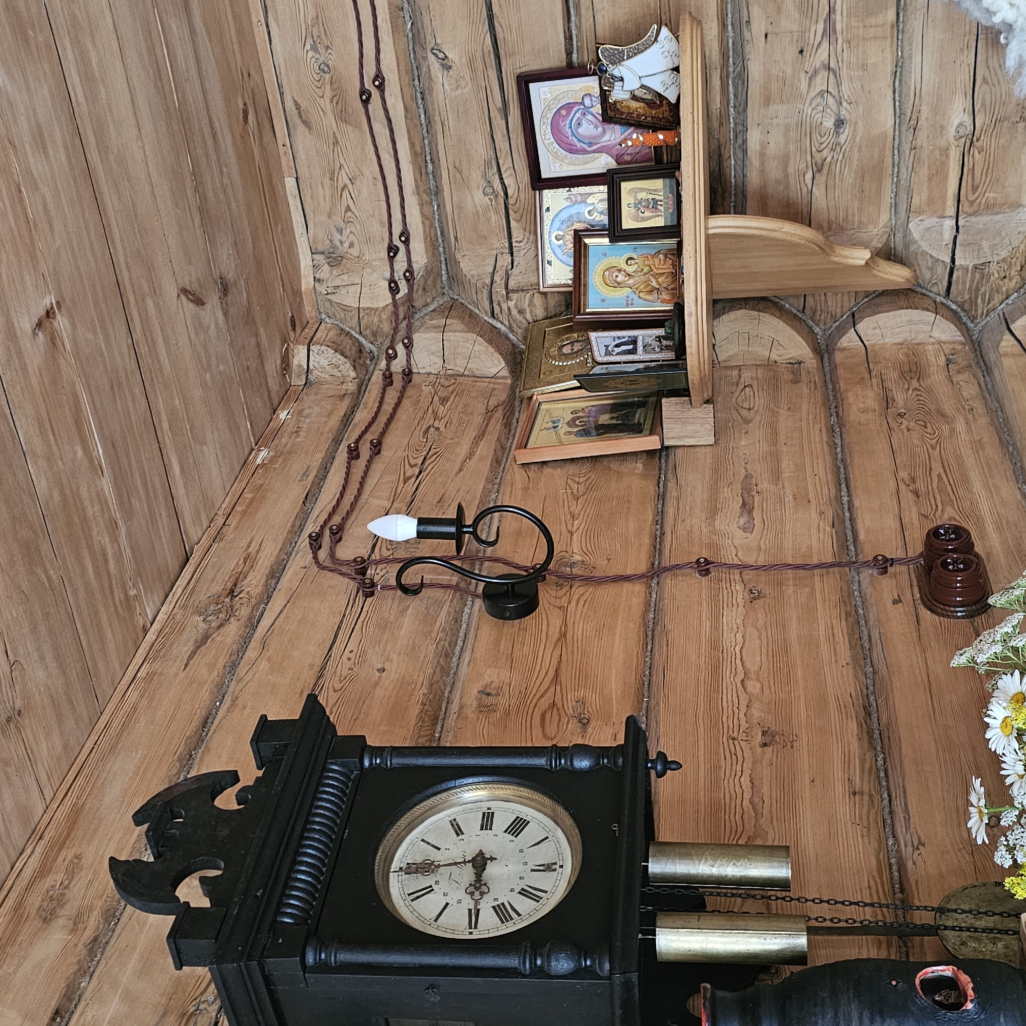 This image depicts an interior scene with rustic wooden walls and flooring, suggesting a dwelling in Vybolovo, Russia. The primary subjects are a collection of religious icons displayed on a wooden shelf, a black grandfather clock, and an antique electric lamp.
The icons, varying in size and framing, are arranged on a light-colored wooden shelf mounted on the wall. They appear to be traditional Orthodox Christian images, with depictions of figures like the Virgin Mary and other saints. Some icons are framed in darker wood, while others are presented more simply. An angelic ornament is attached to the top of the shelf.
A black grandfather clock, with an ornate carved piece on its left side, dominates the lower part of the image. Its face shows Roman numerals and intricate hands, indicating the time is approximately 3:15. Visible on the right side of the clock are two brass weights and chains, suggesting it is a pendulum clock that requires winding.
An old-fashioned electric lamp with a black metal fixture and a white bulb is attached to the wall, connected by a visible, braided wire. This wire runs along the wooden wall and floor, supported by small ceramic insulators. Two similar ceramic electrical outlets are also visible on the wall.
In the lower right corner, a small arrangement of white daisies and other small white flowers adds a touch of natural color to the scene. The lighting suggests it is daytime, with natural light filtering into the room. The overall atmosphere is one of a traditional, perhaps somewhat aged, living space. No visible text is present in the image.