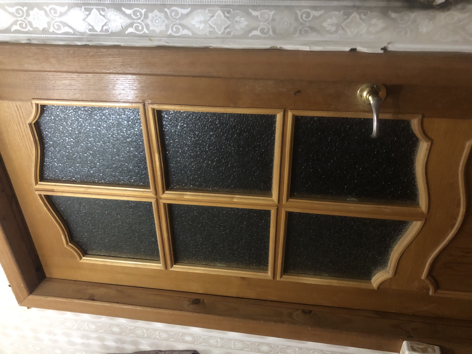 This image shows a close-up, top-down view of a wooden door with glass inserts. The door appears to be part of a home in a settlement in Russia. The glass panes are textured and divided into a 2x3 grid, with decorative wooden trim around each pane. A brass door handle is visible on the right side of the door. Above the door, there is a strip of wallpaper with an ornate, repeating pattern. The overall scene suggests an interior setting, possibly a hallway or entrance. There is no visible text or indication of time of day or weather. The image focuses on the details of the door's construction and design.