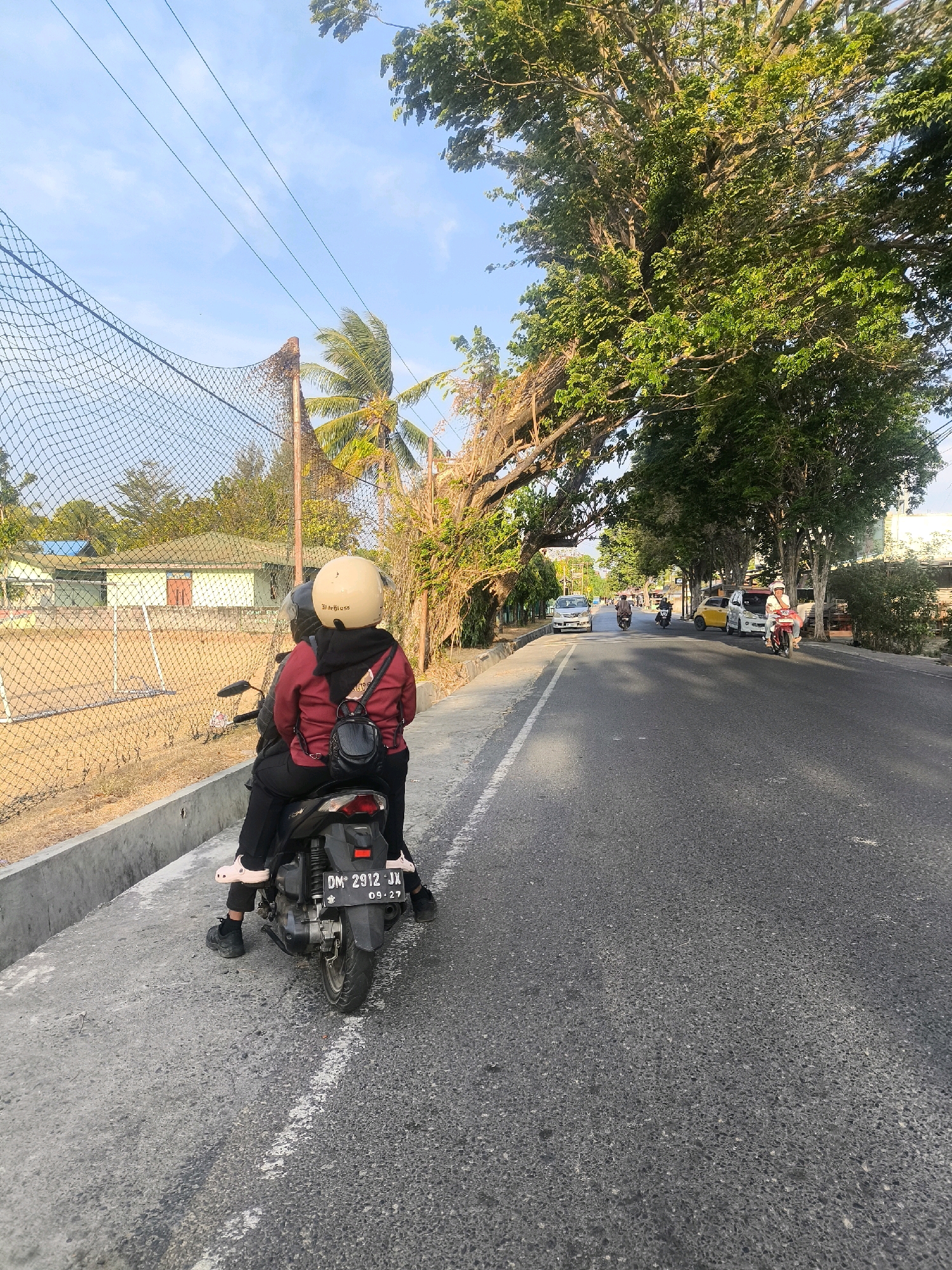 Two individuals are seated on a black scooter, facing away from the camera, on the right side of a paved road. The person in the front wears a light-colored helmet and a red jacket over a black hoodie. The person behind them is wearing a black jacket and black pants. The scooter has a license plate reading "DM 2912 JX 09-27". The road is bordered by a concrete curb and a chain-link fence on the left. Beyond the fence, there is a sandy area with some low buildings and trees. On the right side of the road, a canopy of large trees with dense green foliage arches over the roadway, with sunlight filtering through the leaves. Further down the road, several other vehicles, including motorcycles and cars, are visible. The sky is clear with a few wispy clouds. The scene appears to be in a tropical urban environment, consistent with Gorontalo, Indonesia, likely during daylight hours.