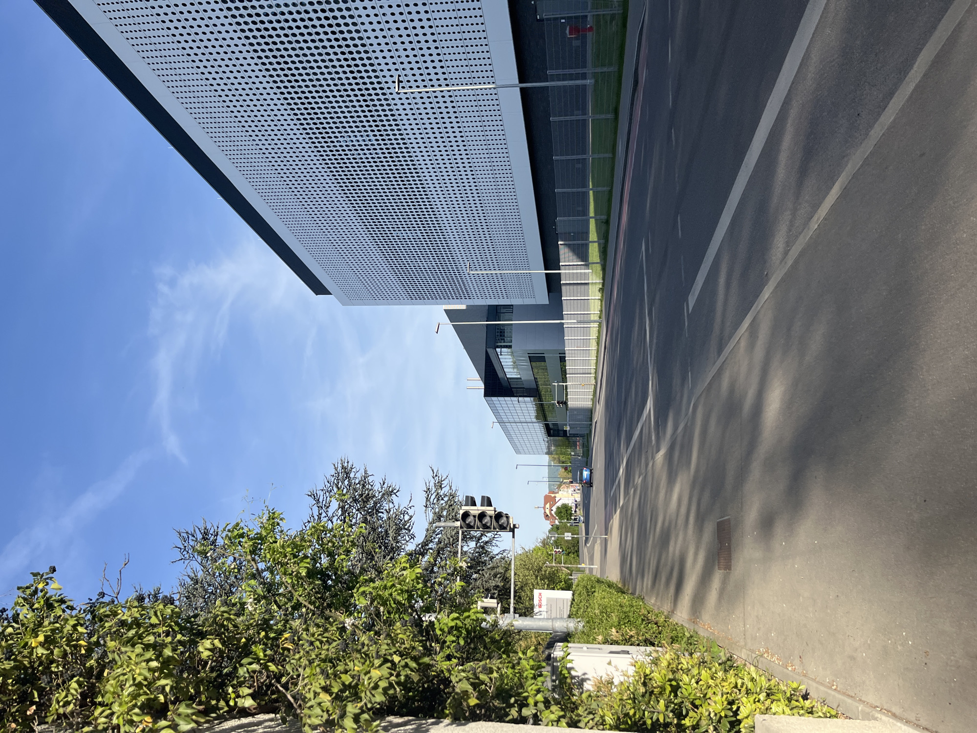 The image captures a street scene in an urban environment. A modern building with a perforated facade dominates the left side of the frame, contrasting with the clear blue sky visible above. The street runs along the right, marked with painted lane lines and bordered by greenery. In the middle ground, a traffic light is partially obscured by foliage. Further down the street, another building is visible, along with a glimpse of distant houses or buildings. A car is also visible further down the street. The environment is sunny, casting shadows across the street and adding depth to the scene.
