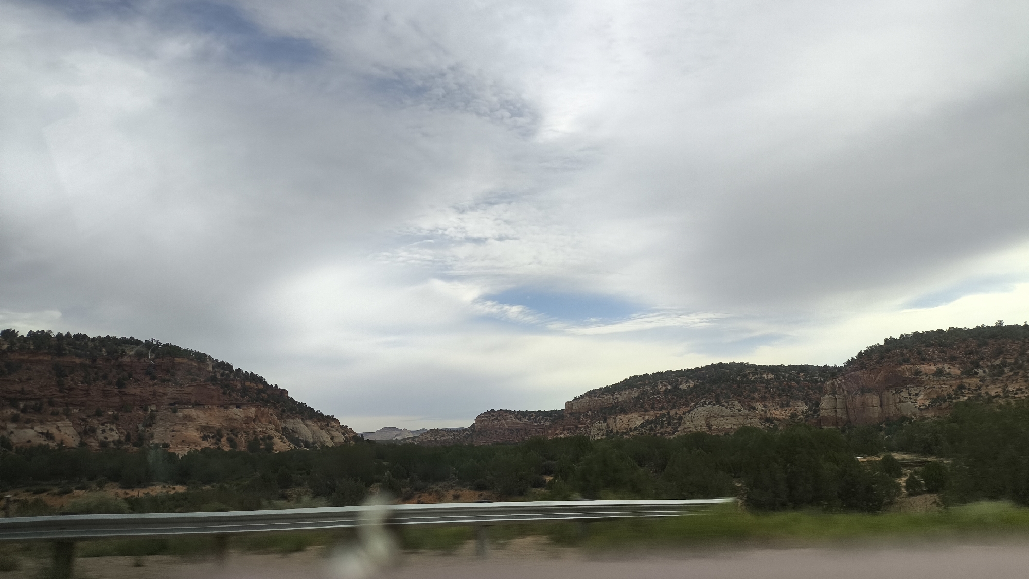 The image shows a vast, arid landscape with layered rock formations and a dense line of green trees in the foreground. The sky is filled with dramatic, heavy clouds, with patches of blue visible. A metal guardrail runs horizontally across the bottom of the frame, suggesting the photo was taken from a vehicle traveling along a road. The overall impression is one of rugged natural beauty. The location is identified as Kanab, United States, a city known for its proximity to numerous national parks and its distinctive desert terrain.