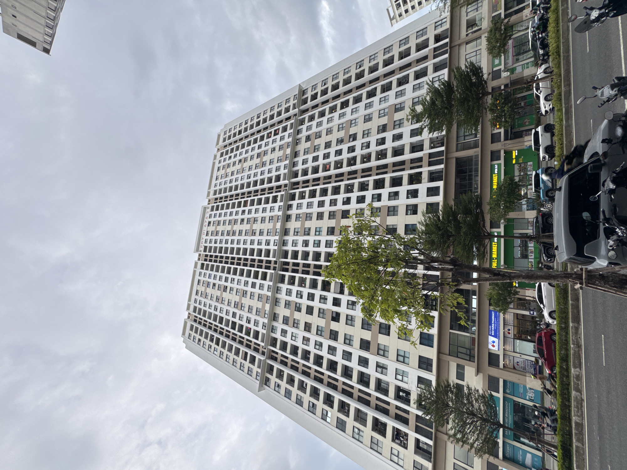 The image captures a wide-angle view of a modern, tall apartment building in Da Nang, Vietnam, under a cloudy sky. The building, a prominent high-rise, features a repetitive pattern of balconies and windows across its numerous floors. The ground level of the building houses several commercial establishments, including a pharmacy with a blue sign reading "NHÀ THUỐC BẢO HUNG PHARMACY" and a green sign indicating "FULL-MARKET OPEN 24/7".
In front of the building, a street is visible with several vehicles, including cars and motorbikes. Some people are seen interacting with these vehicles, suggesting a busy urban environment. The presence of trees lining the street adds a touch of greenery to the scene. The perspective of the photo is from a low angle, looking up towards the immense structure, emphasizing its scale and architectural design. The overall impression is one of urban development and daily life unfolding in a Vietnamese city.
