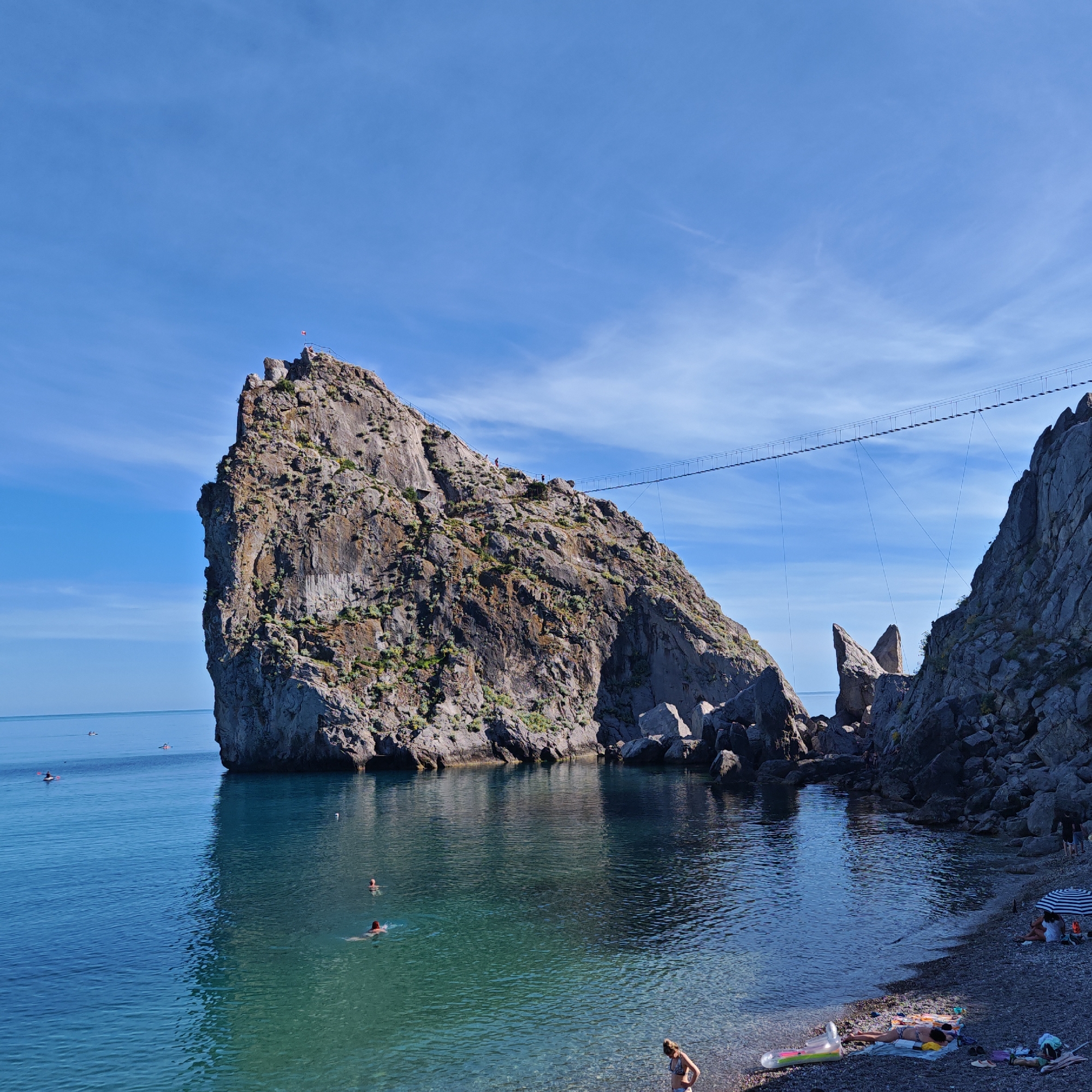 This image depicts a scenic coastal view in Simeiz, Ukraine, with a prominent rock formation jutting into the sea. The rock is steep and craggy, with sparse vegetation growing on its surface. A suspension bridge spans across the gap between this rock and another smaller rock formation in the distance, suggesting a popular tourist or recreational area.
The water is a clear, calm turquoise, with several people swimming and kayaking in the sea. On the pebbled shore in the foreground, several individuals are relaxing, some under a beach umbrella, and others sunbathing on towels. The sky is a bright, clear blue with a few wispy clouds. The overall impression is one of a tranquil summer day at a beautiful seaside location.