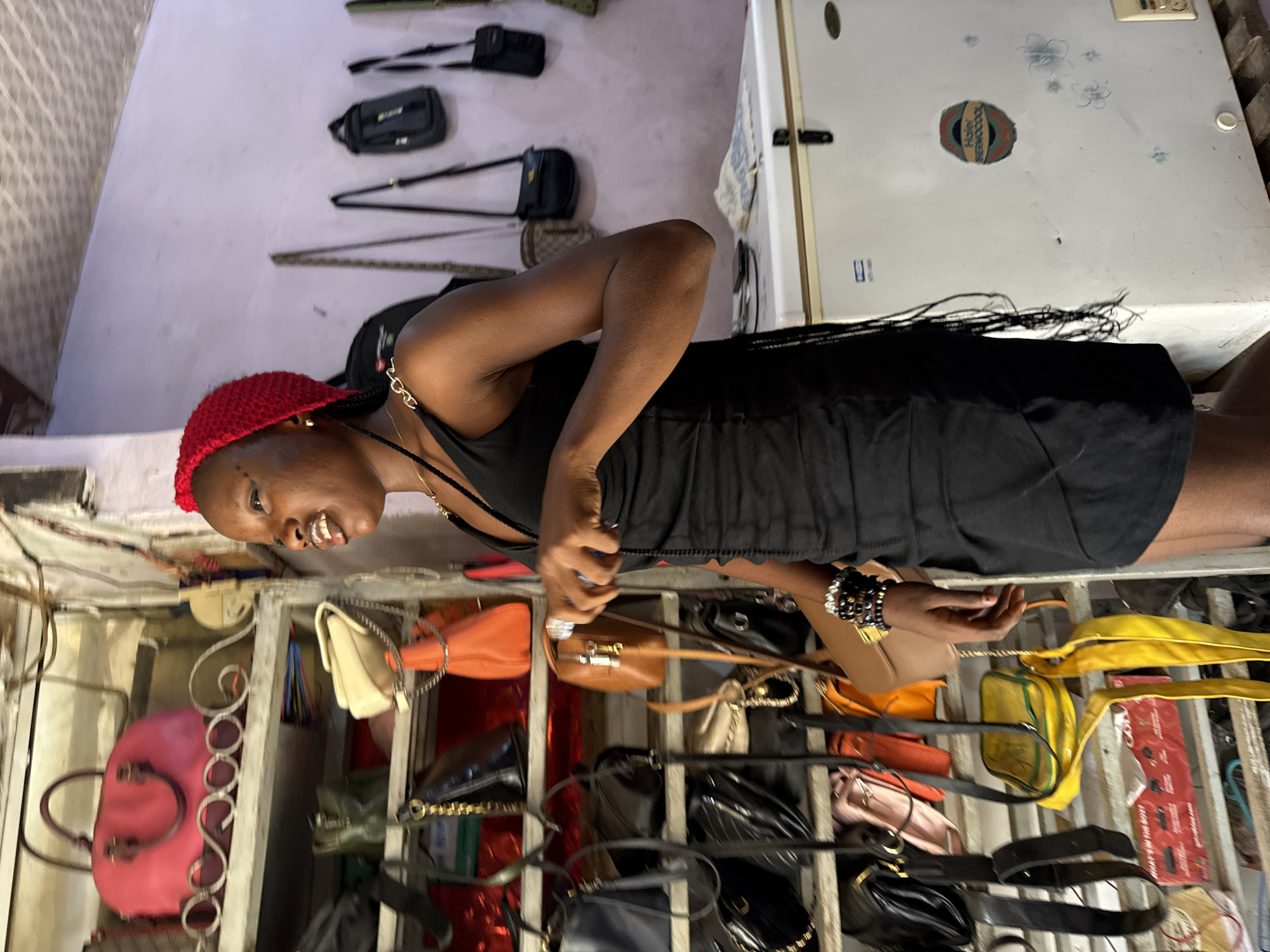 A woman with a red head covering, wearing a black dress, is standing in front of a rack of handbags in a shop in Abuja, Nigeria. The handbags are various colors, including pink, orange, yellow, and black. Behind the woman is a white refrigerator. A rack of other bags and equipment is visible hanging on the wall.